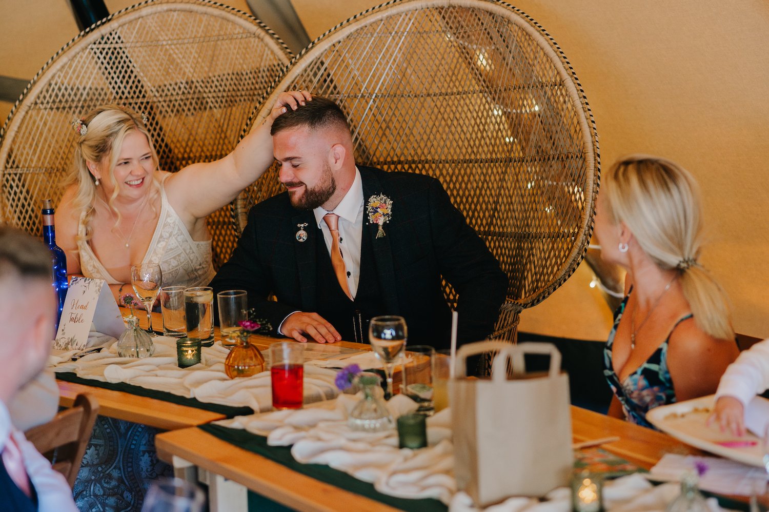 A woman playfully touches a man's head as they sit at a decorated table during a celebration. The woman has blonde hair styled in waves, and the man has short hair and a beard, wearing a dark blazer with a floral boutonniere. Other women and table se