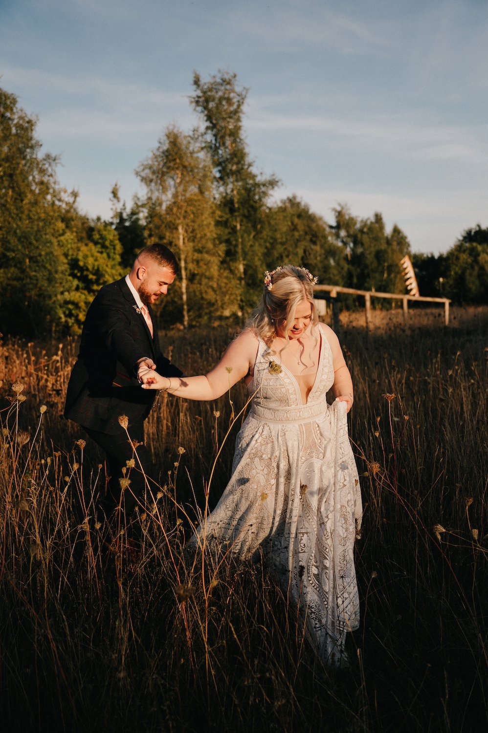 A bride and groom walking through a field at sunset, holding hands, with the bride smiling and wearing a detailed white wedding dress and floral crown, while the groom is dressed in a dark suit.