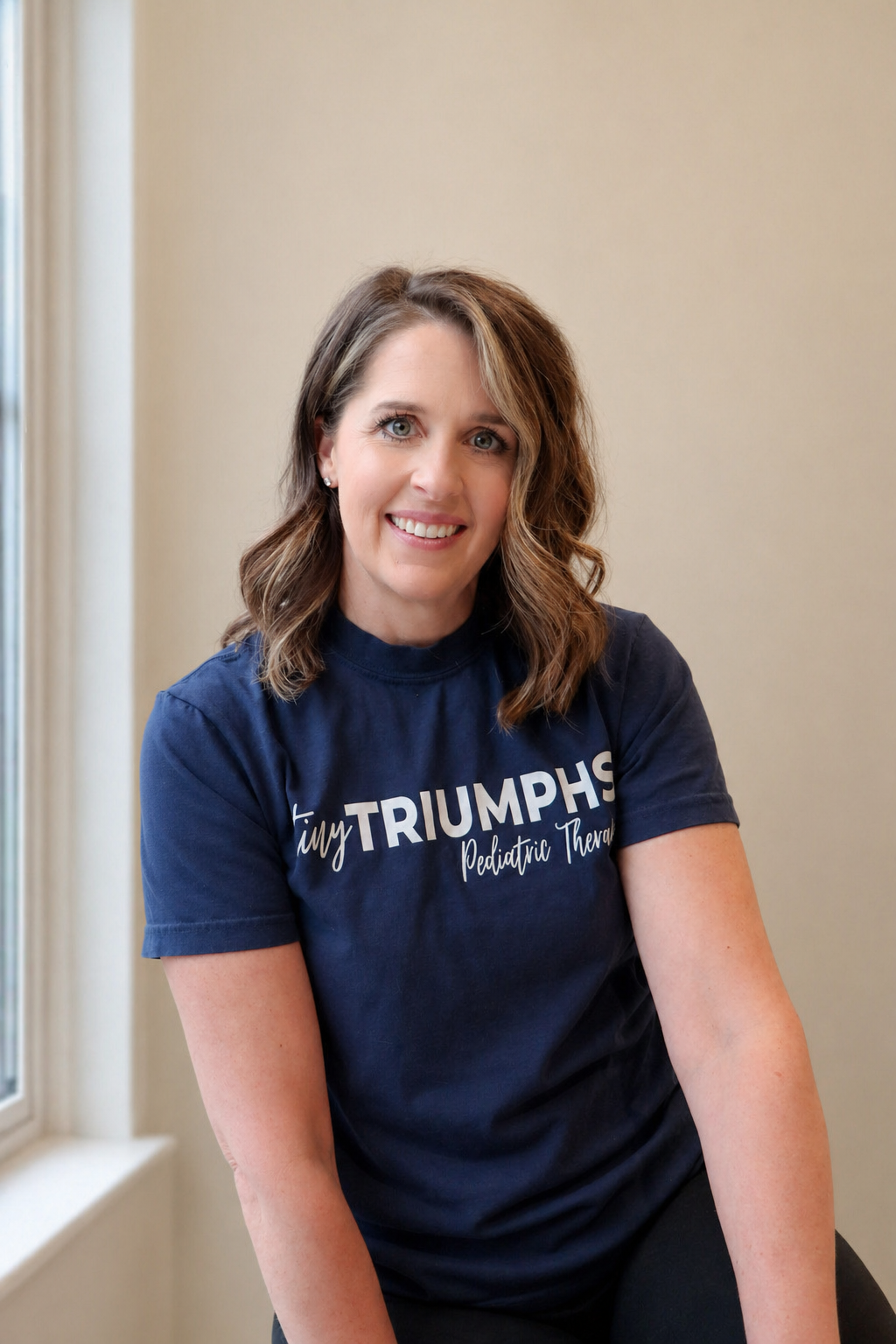 A woman with medium-length brown hair, blue eyes, and a bright smile, wearing a navy blue T-shirt with white text that reads 'tiny TRIUMPH'S Pediatric Therapy,' sitting indoors near a window.