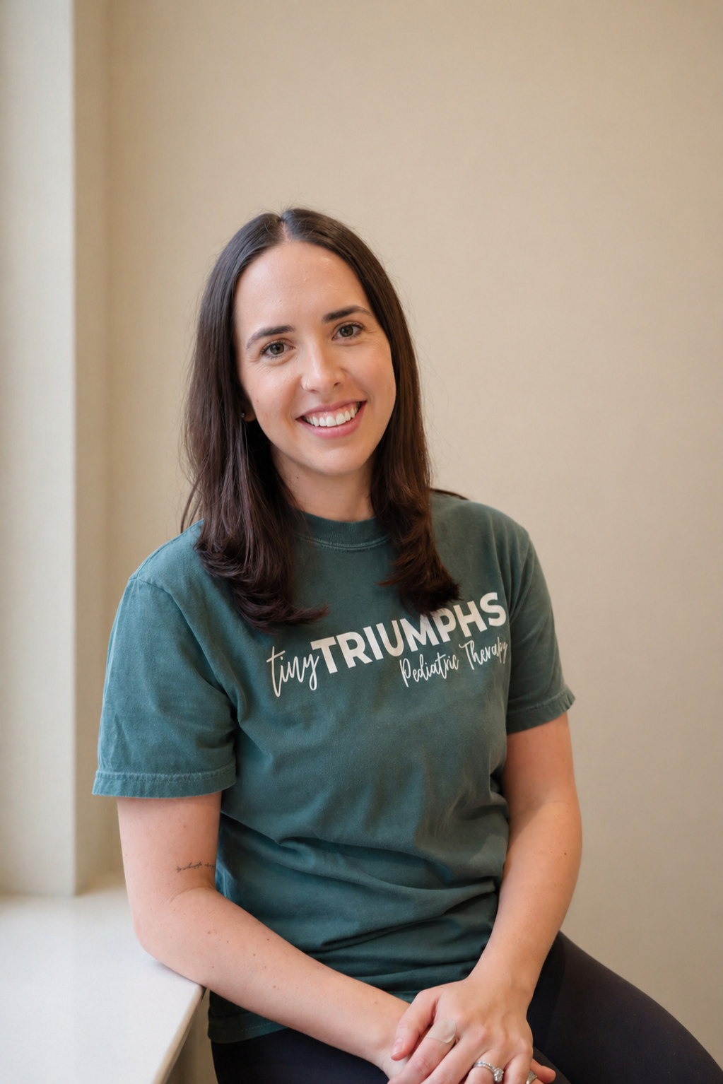A smiling young woman with shoulder-length brown hair, wearing a teal T-shirt with white text that reads 'tinyTRIUMphs Pediatric Therapy,' sitting against a beige wall.