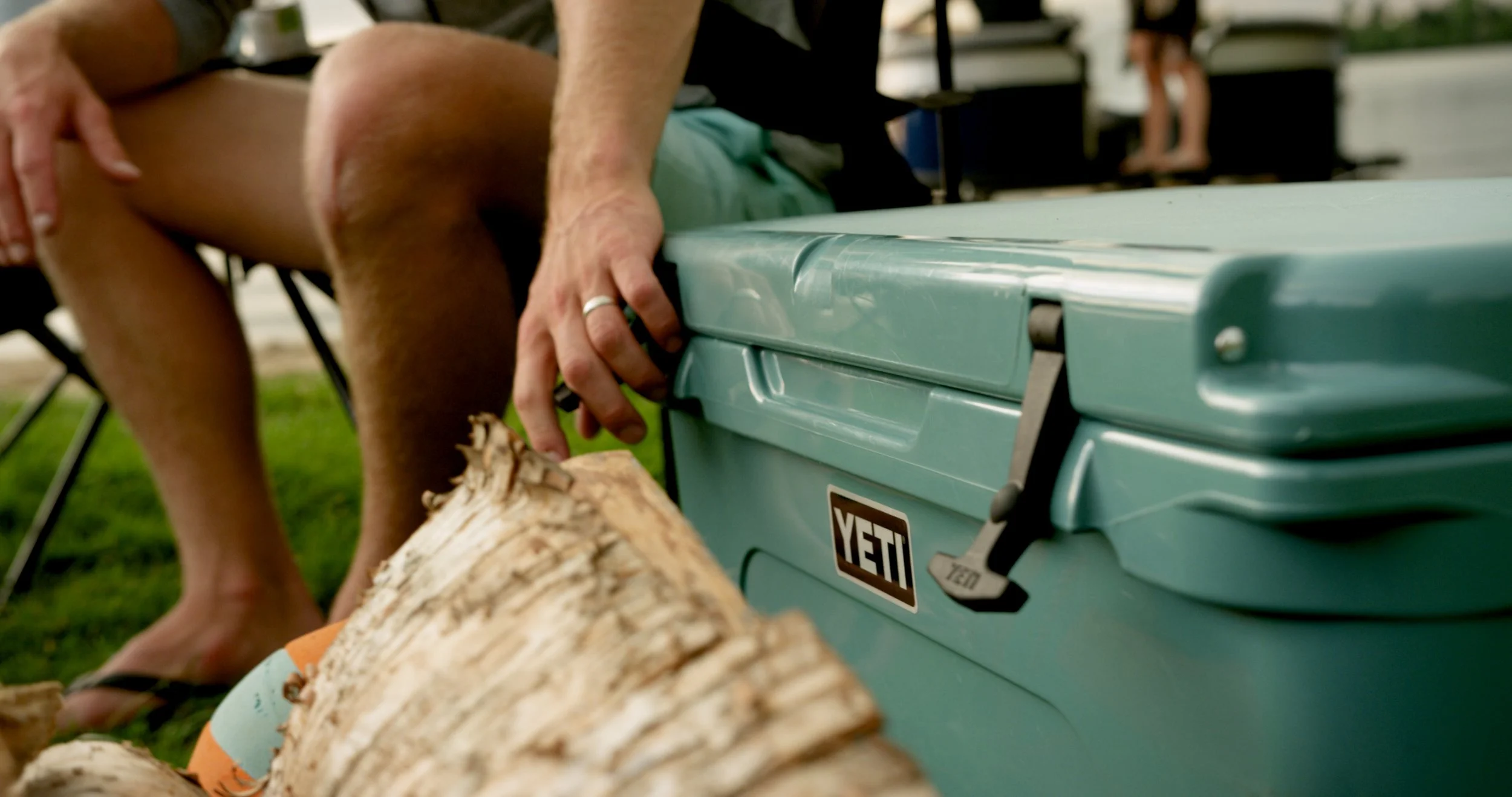 Close-up of a person's hand turning a latch on a light blue YETI cooler, with logs and grass nearby.