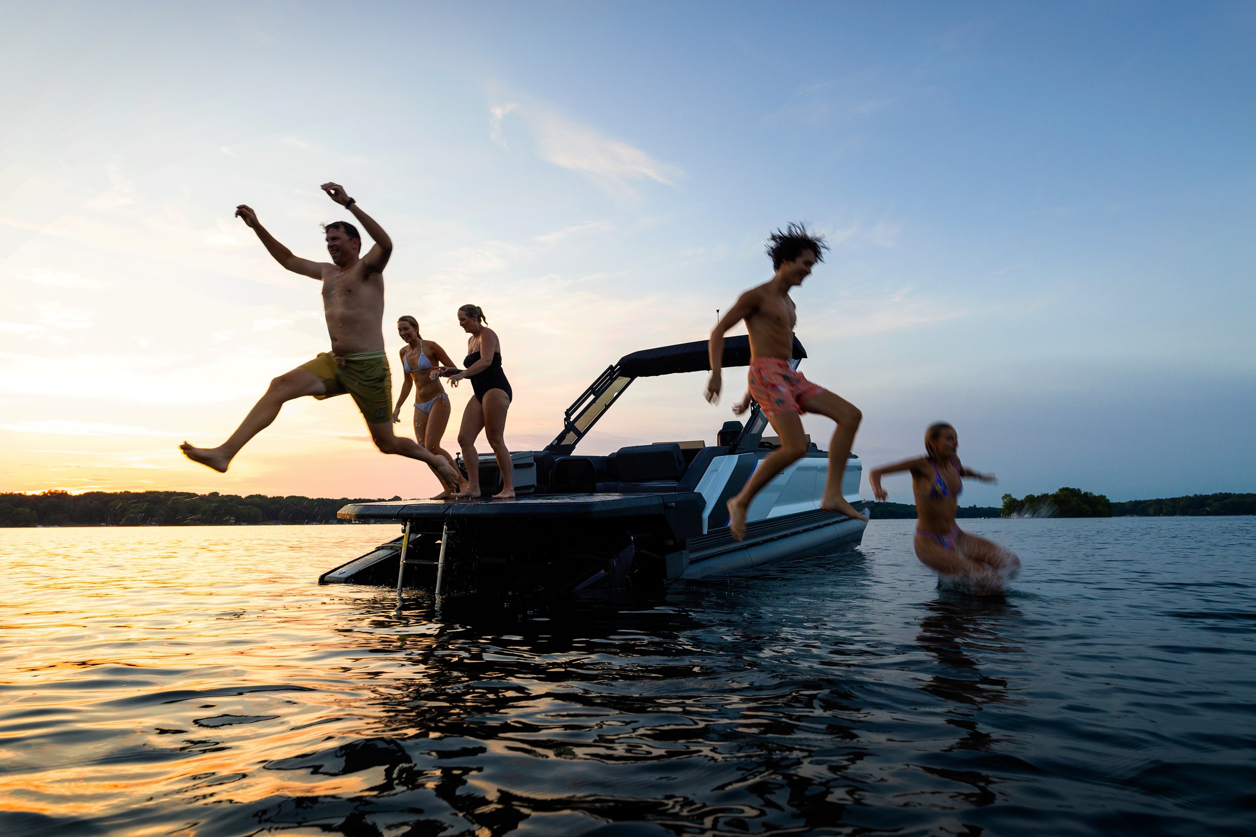 People enjoying a boat on a lake at sunset, jumping into the water, wearing swimsuits, with a clear sky in the background.
