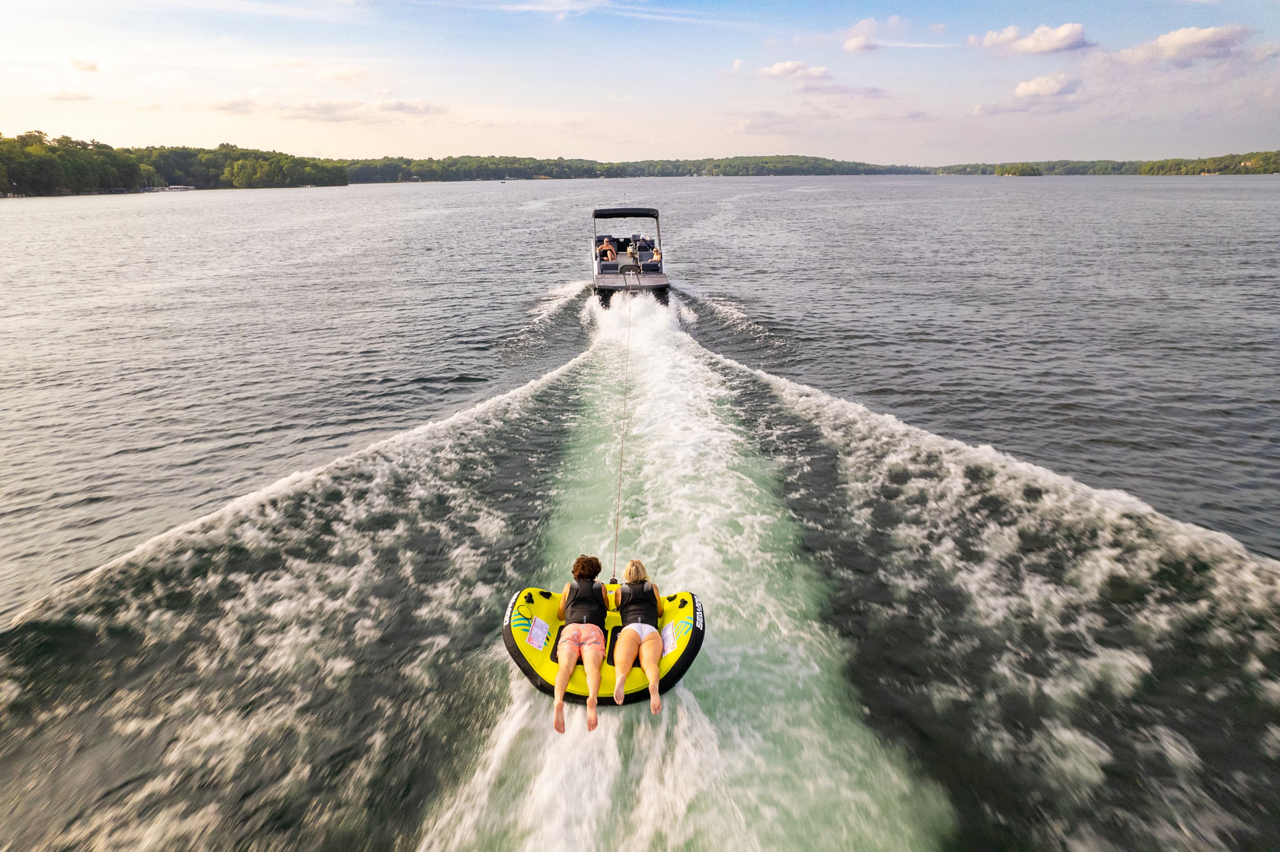 People riding an inflatable tube being pulled by a boat on a lake, with a child in a life jacket hanging on the tube, and trees along the shoreline in the background.