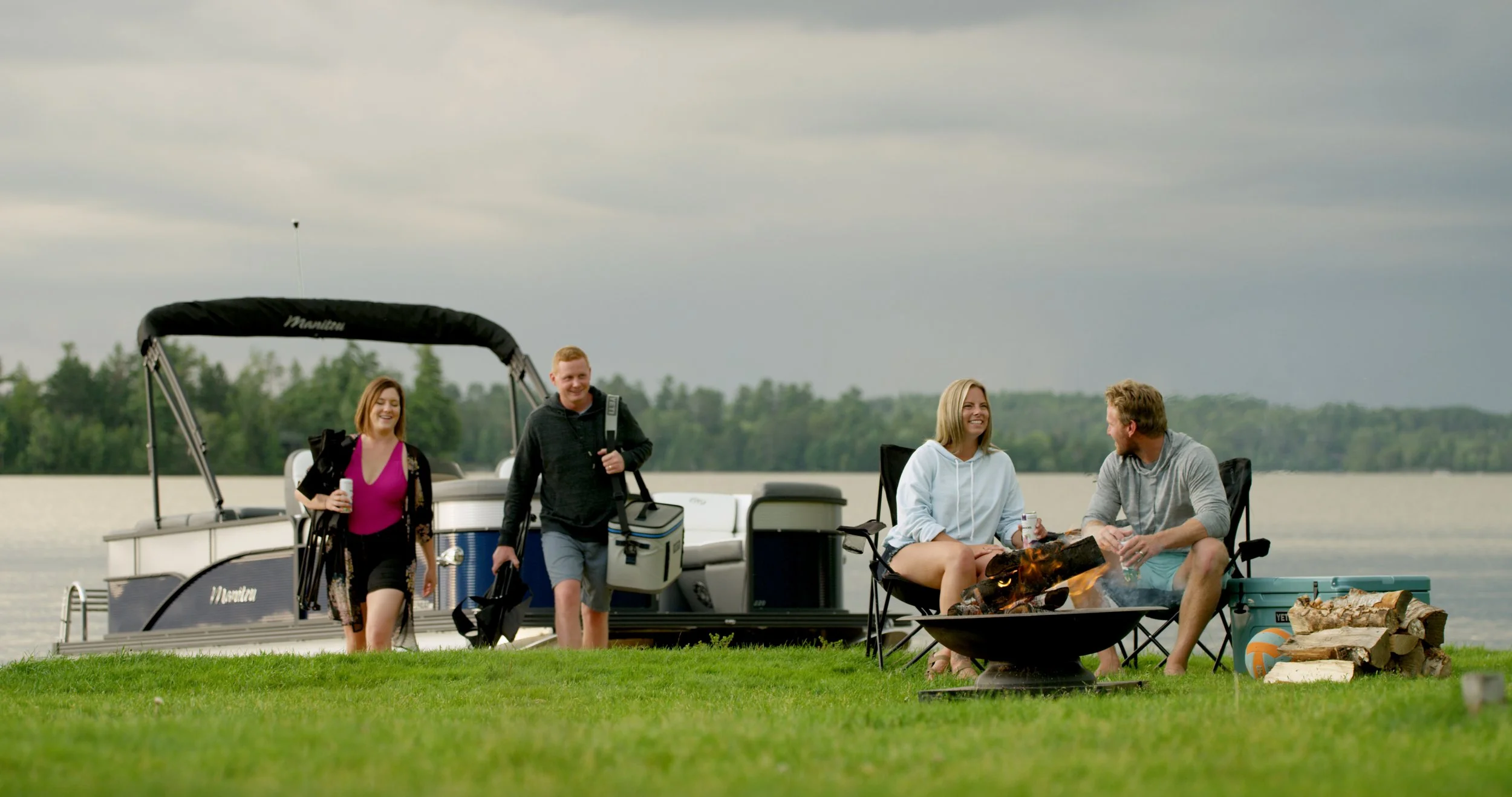 Four people enjoying a boat trip and outdoor barbecue by a lake on an overcast day. Two women sit by a fire pit, while two men walk toward them holding drinks and a cooler; a boat is docked nearby.