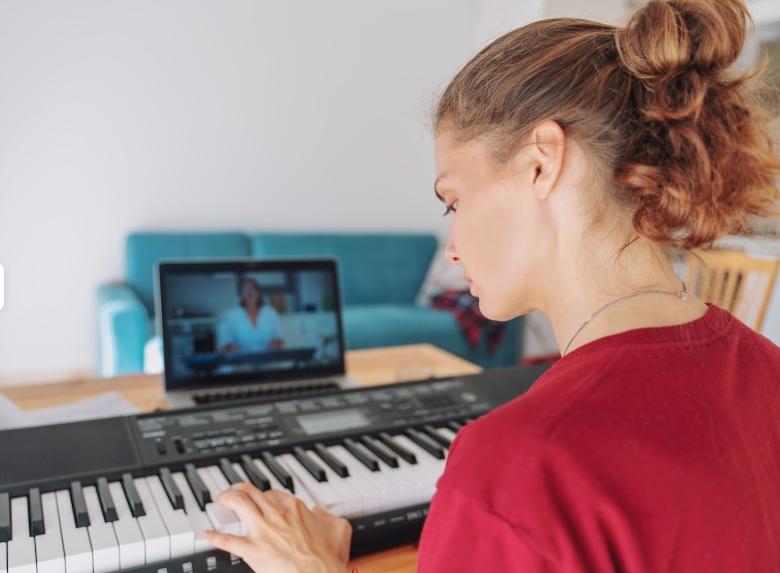 Young woman with curly hair in a bun practicing piano while on a video call on her laptop in a living room.