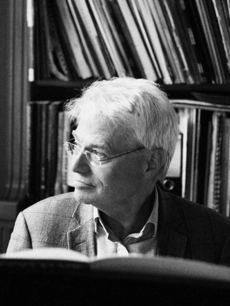 A black and white photo of an elderly man with a slight smile, sitting in front of a bookshelf.