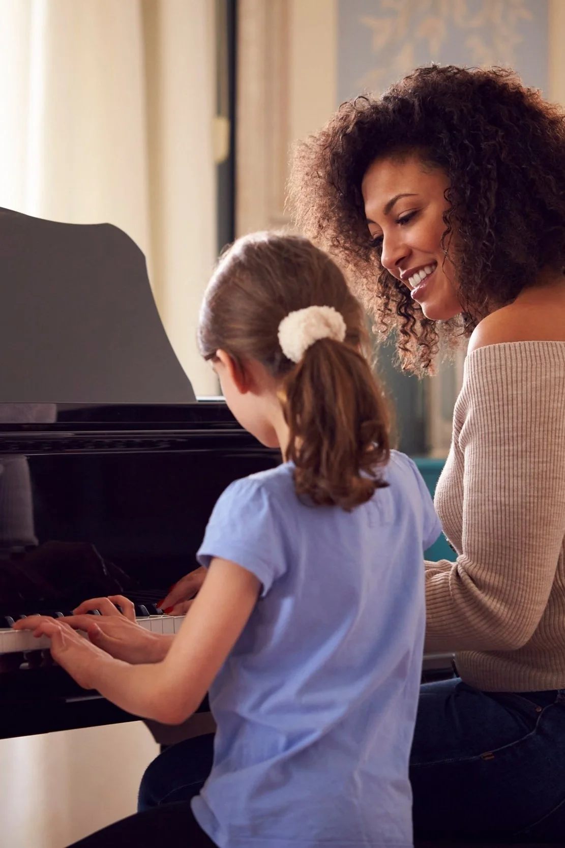 A woman and a young girl sit at a piano, with the woman smiling and guiding the girl as she plays.