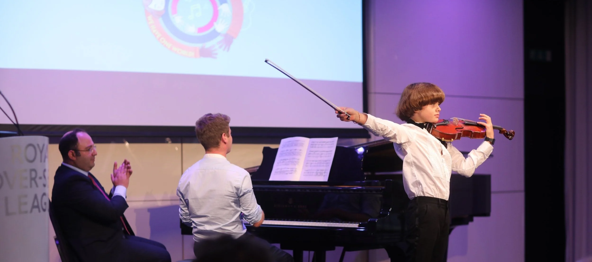 A young boy performing a violin piece with piano accompaniment at a concert.