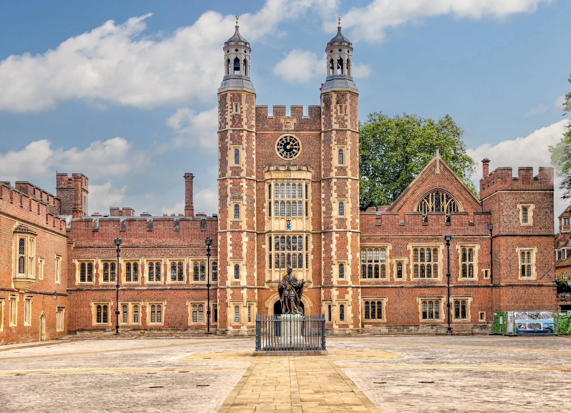 A historic brick building with a central clock tower topped with a weather vane, surrounded by a courtyard with a statue of a person in medieval attire.