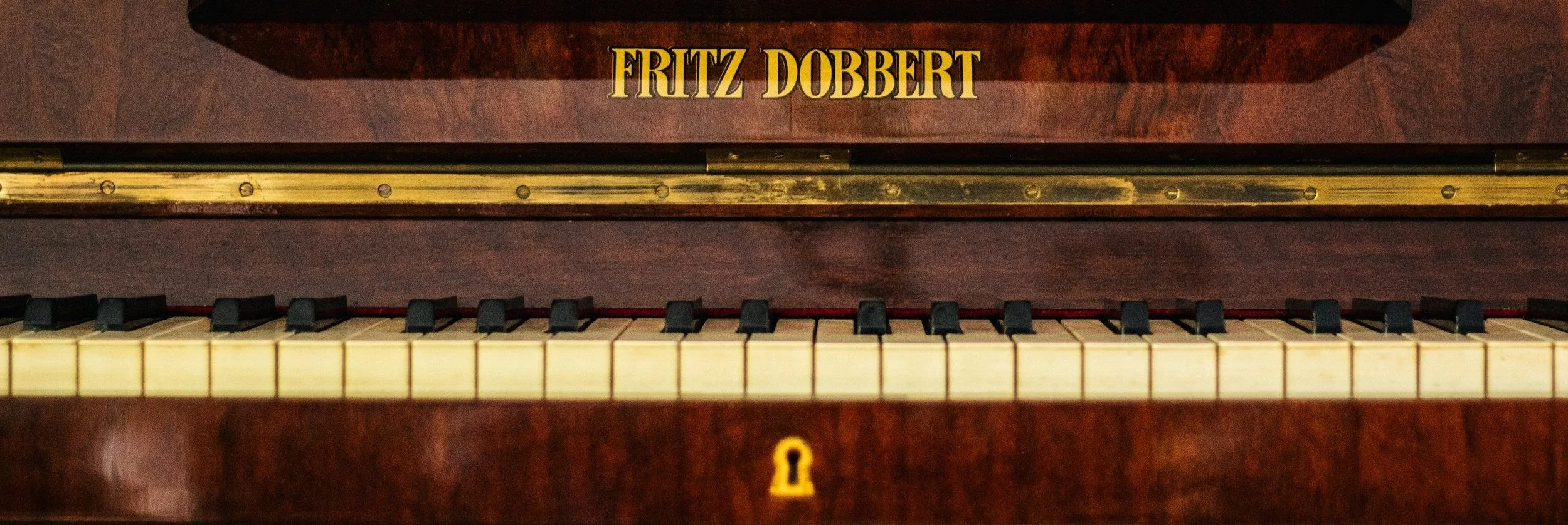 Top view of a vintage piano with a wood finish, black and white keys, and a keyhole, with a brass hinge and a sign that reads "Fritz Dobbert".