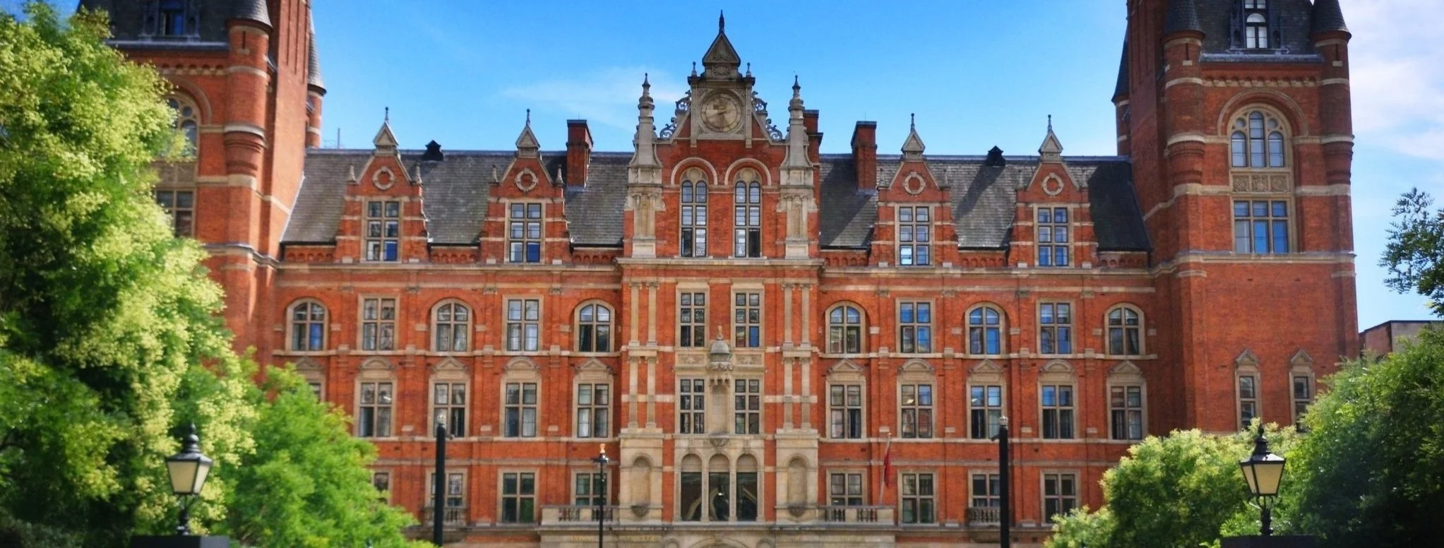 Historic red brick building with ornate architecture and multiple windows, surrounded by green trees under a blue sky.