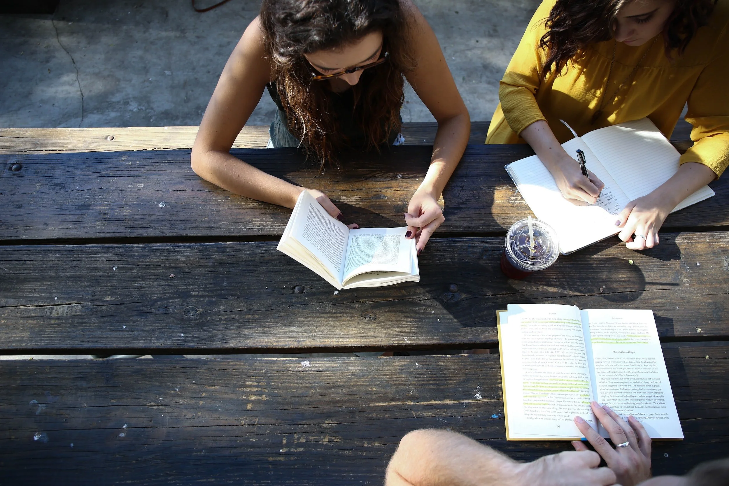 Two women sitting at a dark wooden table outdoors, reading books. One woman in a black top is holding a small book, the other in a yellow top is writing in a notebook. There is a drink with a straw on the table, and a third open book in the foreground.