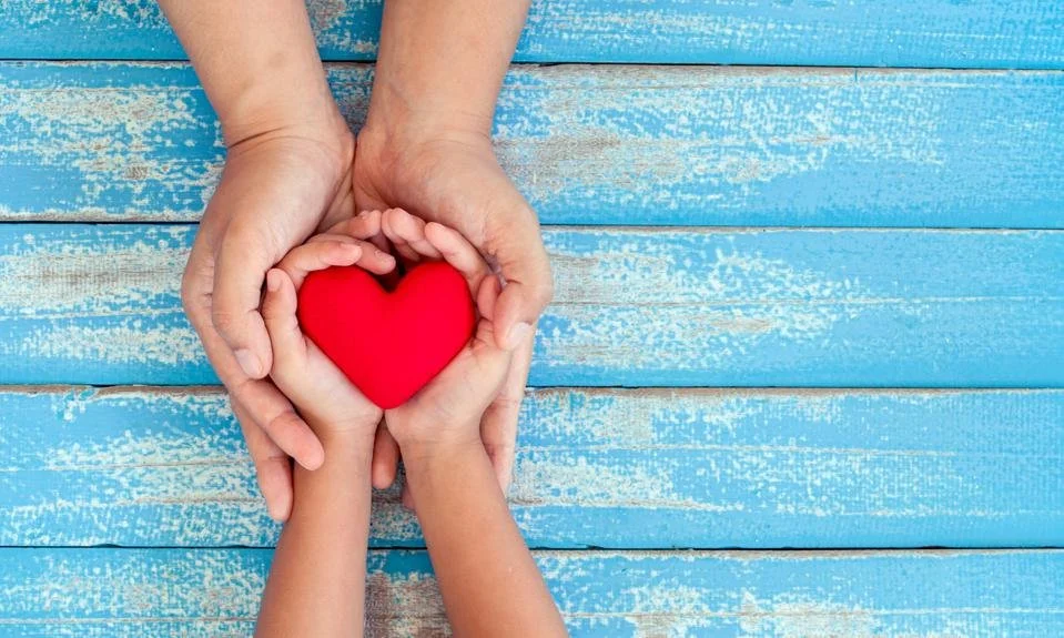 Two adult hands and two child's hands holding a red heart-shaped object over a blue wooden surface.