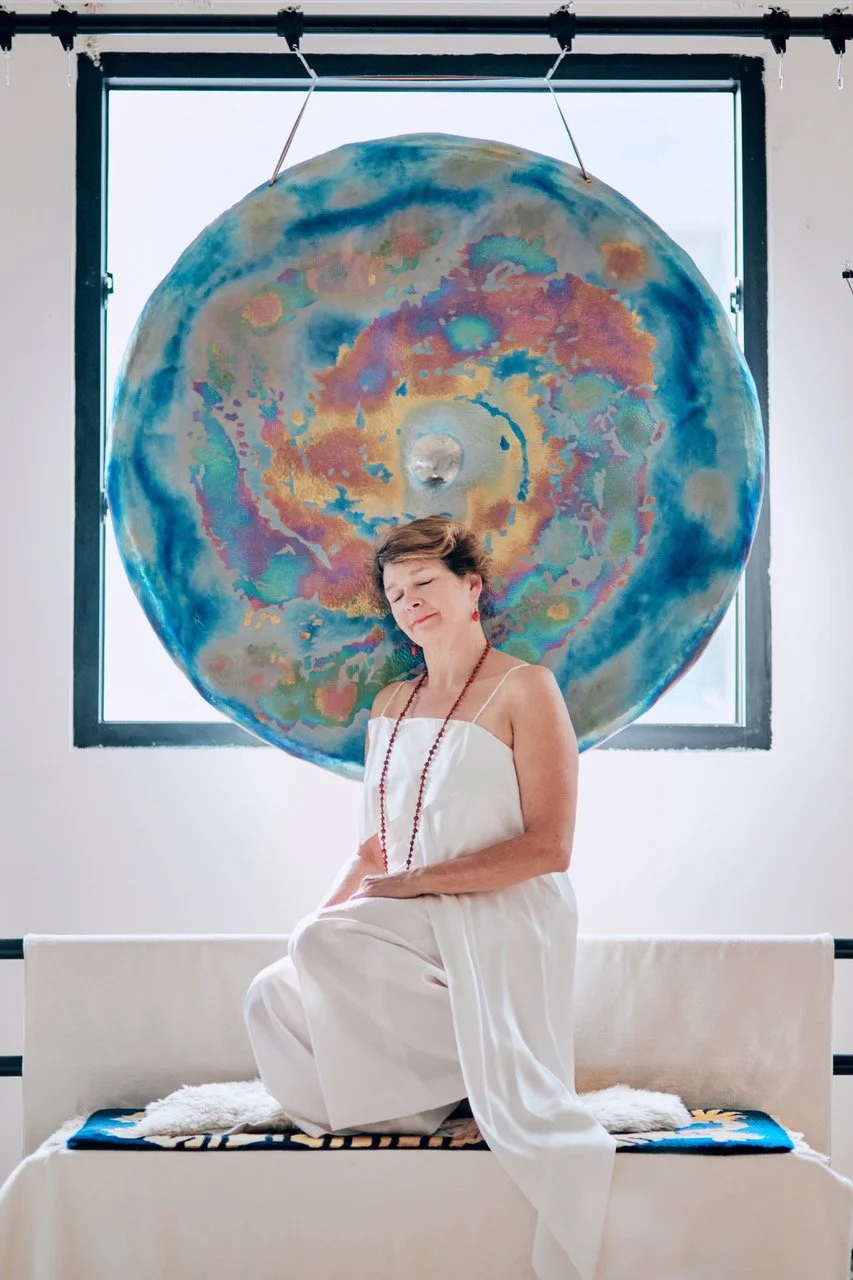 Martha Collard seated in meditation before a large hand-painted gong at Red Doors Studio