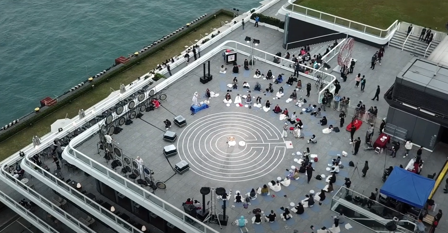Aerial view of a rooftop terrace at Harbour City beside Victoria Harbour, with a labyrinth at its centre, a line of gongs along the railing and guests seated around it.