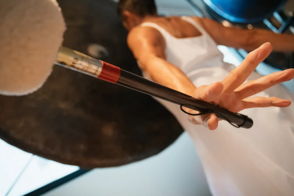 Martha Collard in white, arm outstretched, playing a large gong with a felt mallet.