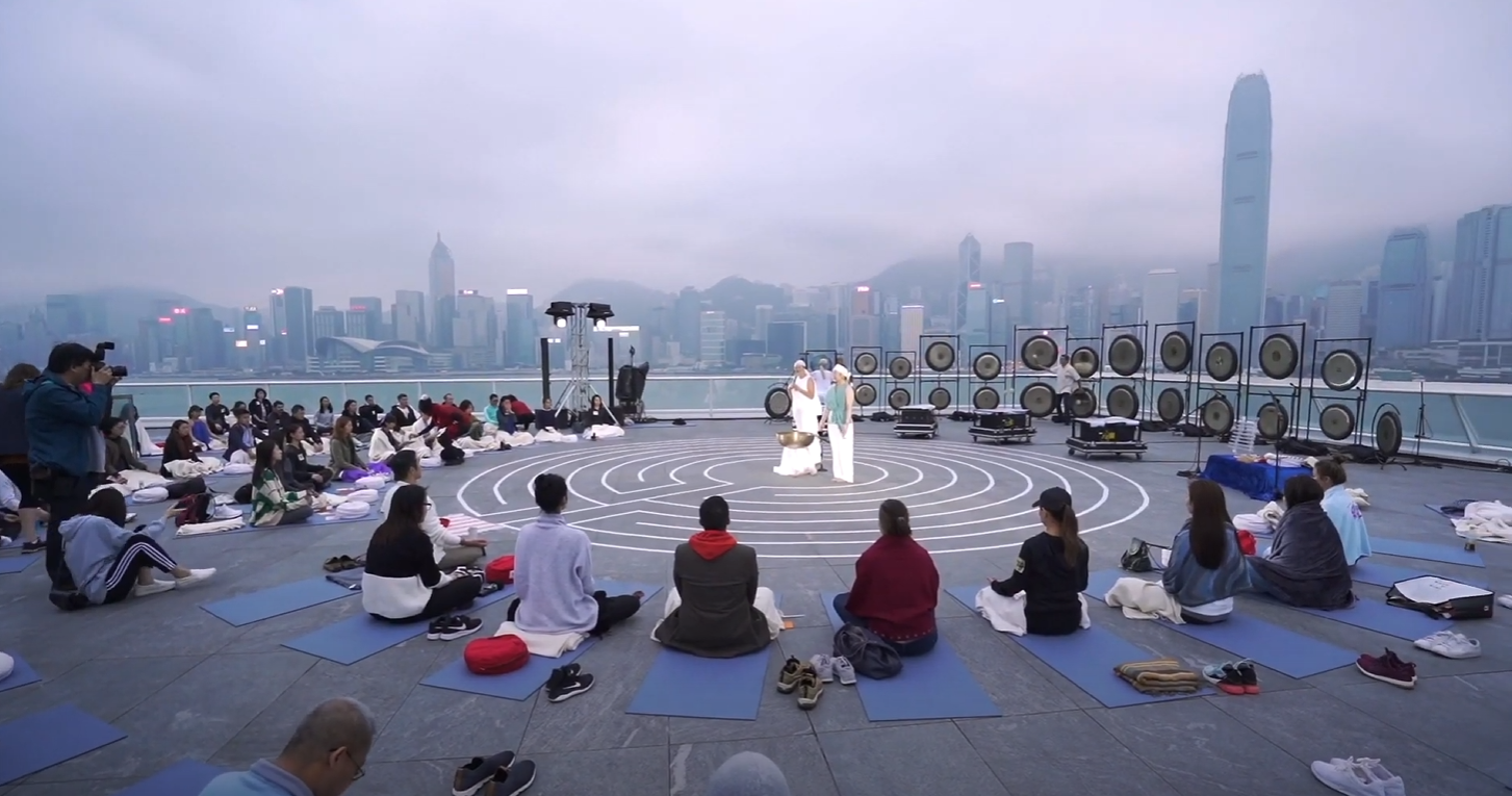 An outdoor session at Harbour City: guests seated around a labyrinth with a wall of gongs behind it and the Hong Kong skyline across the harbour.