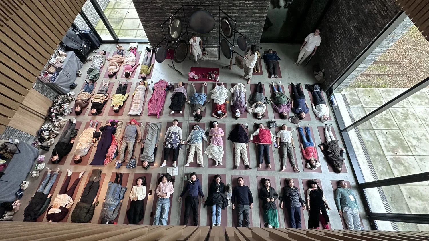 Seen from above at the Sangha Octave At-One International Festival in Suzhou: four rows of people lying on mats in a tall concrete hall, with a rig of gongs at the far end.