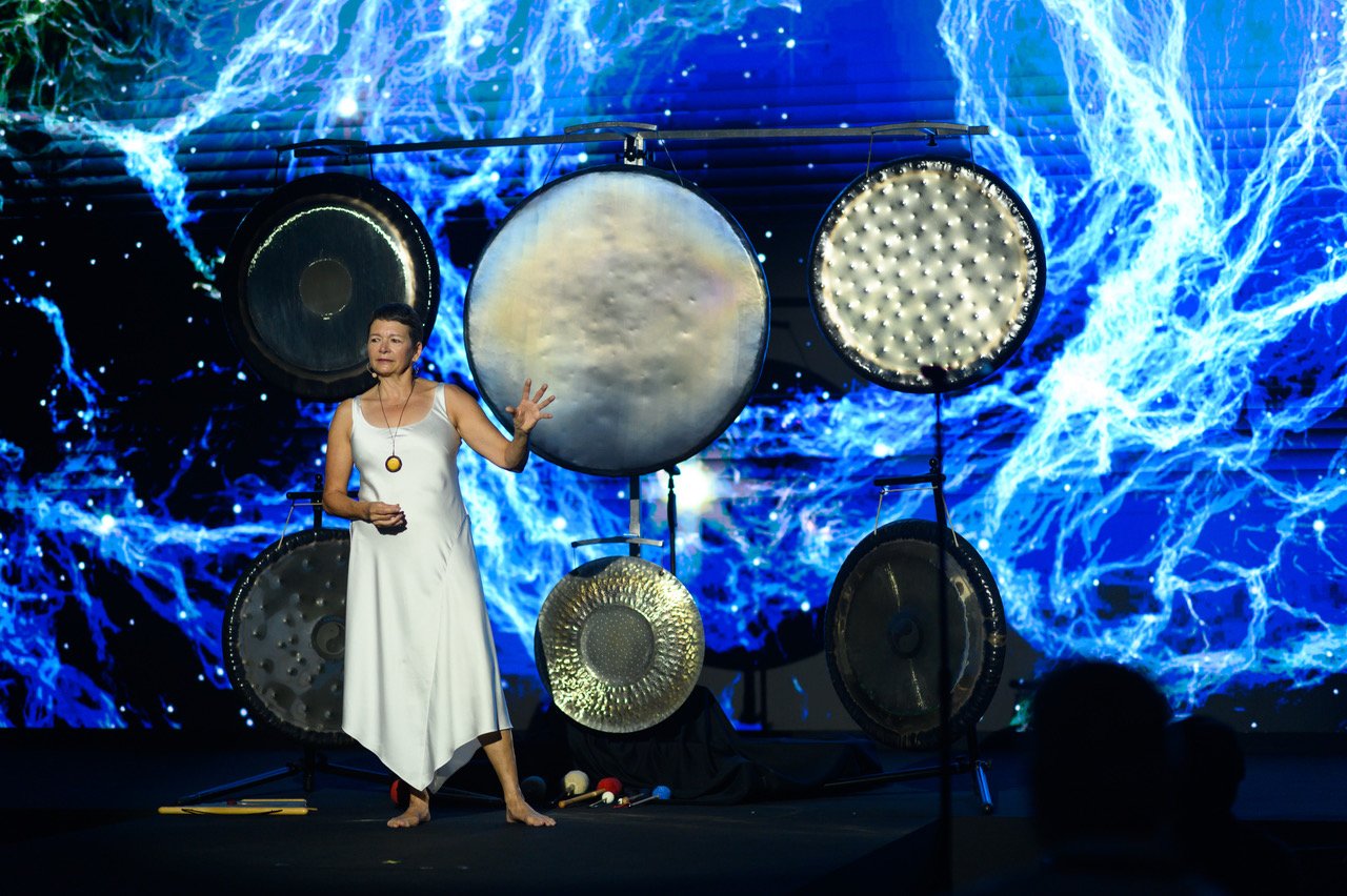 Martha Collard in a white dress on stage, gesturing towards a set of gongs in front of a bright blue screen.