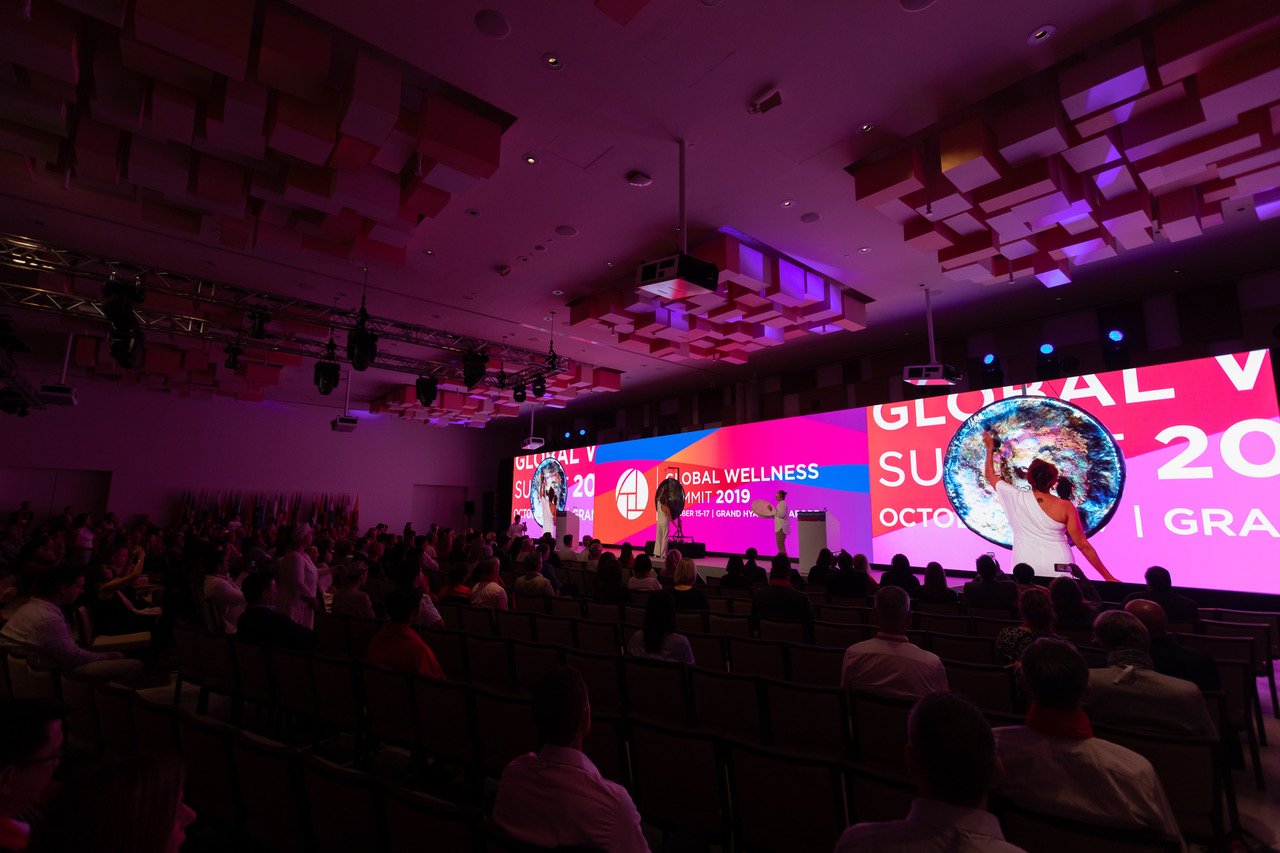 A dark conference hall at the Global Wellness Summit 2019, the audience seated facing a stage where a gong is being played, shown large on the screens either side.