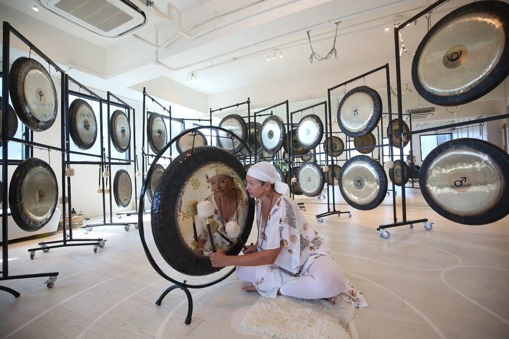 A woman in white kneels to play a gong at Red Doors Studio, surrounded by dozens of gongs on black stands