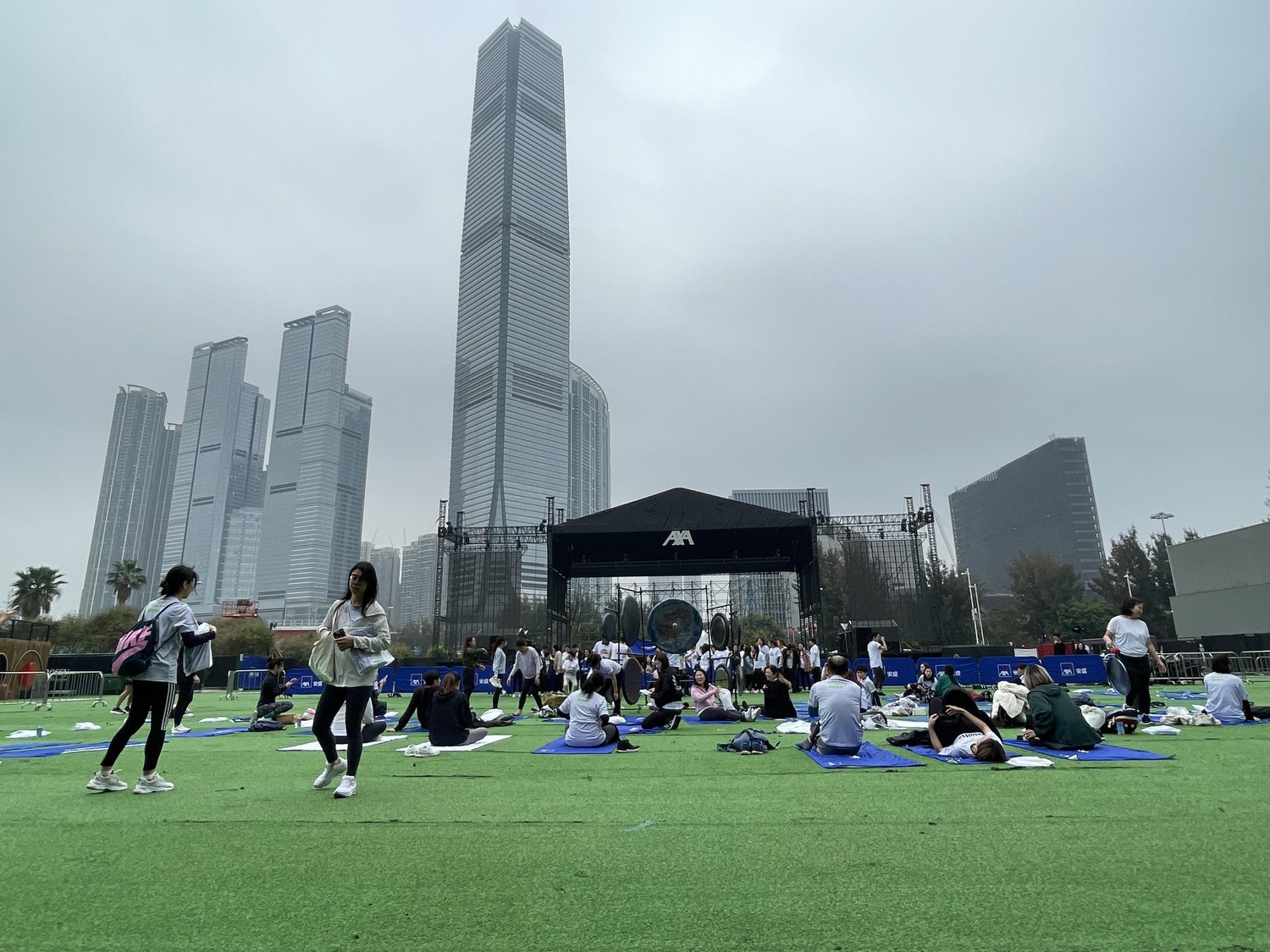 An open-air lawn in West Kowloon with people on mats in front of an AXA-branded stage holding a rig of gongs, the ICC tower behind.