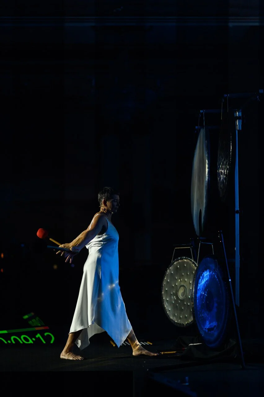 Martha Collard in a white dress on a dark stage, mallets in hand, walking towards a stand of gongs.