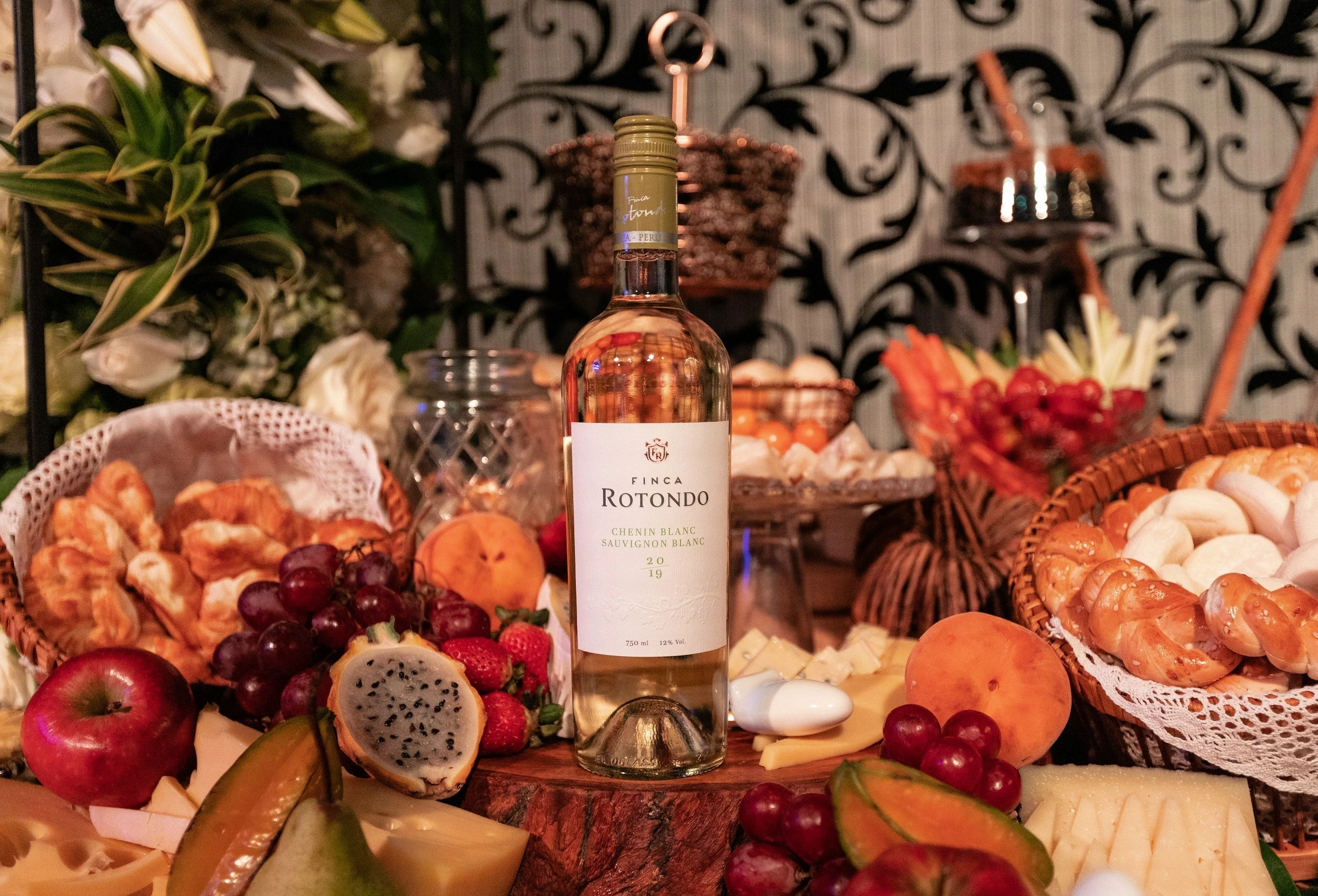 A bottle of Finca Rotondo Chenin Blanc and Sauvignon Blanc wine surrounded by various fruits and cheeses on a festive table.