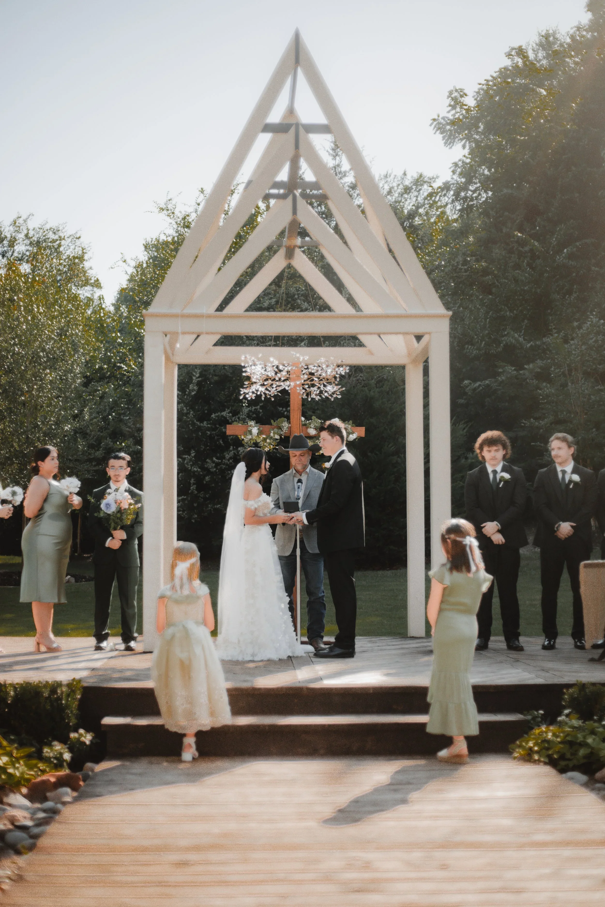 A wedding ceremony is taking place outdoors under a small wooden pavilion with a cross and hanging chandelier, with a group of people dressed formally, including a bride and groom exchanging vows, with bridesmaids and groomsmen nearby, and wedding gu