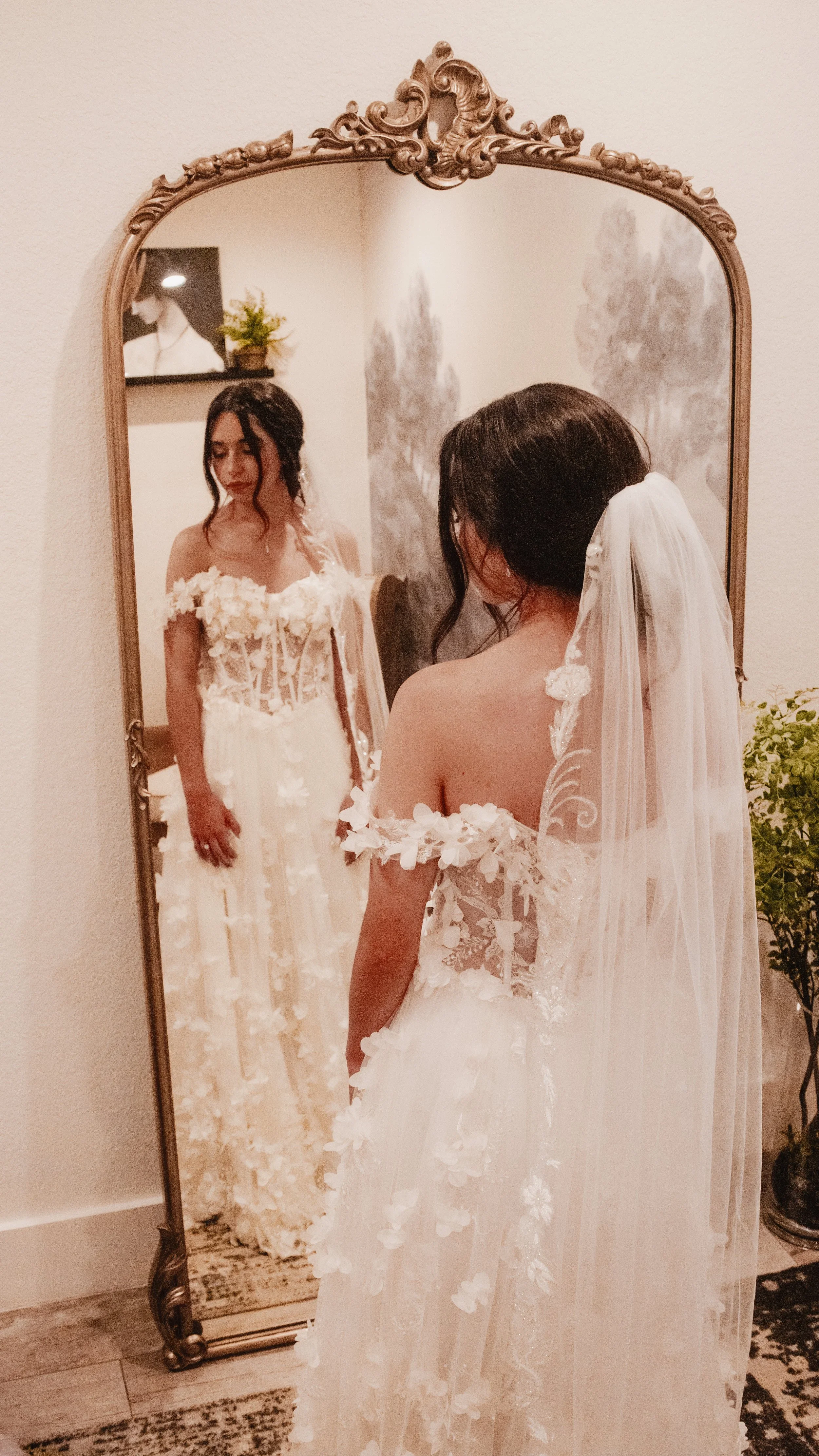 A woman in a wedding dress with floral details is looking at herself in a full-length mirror.