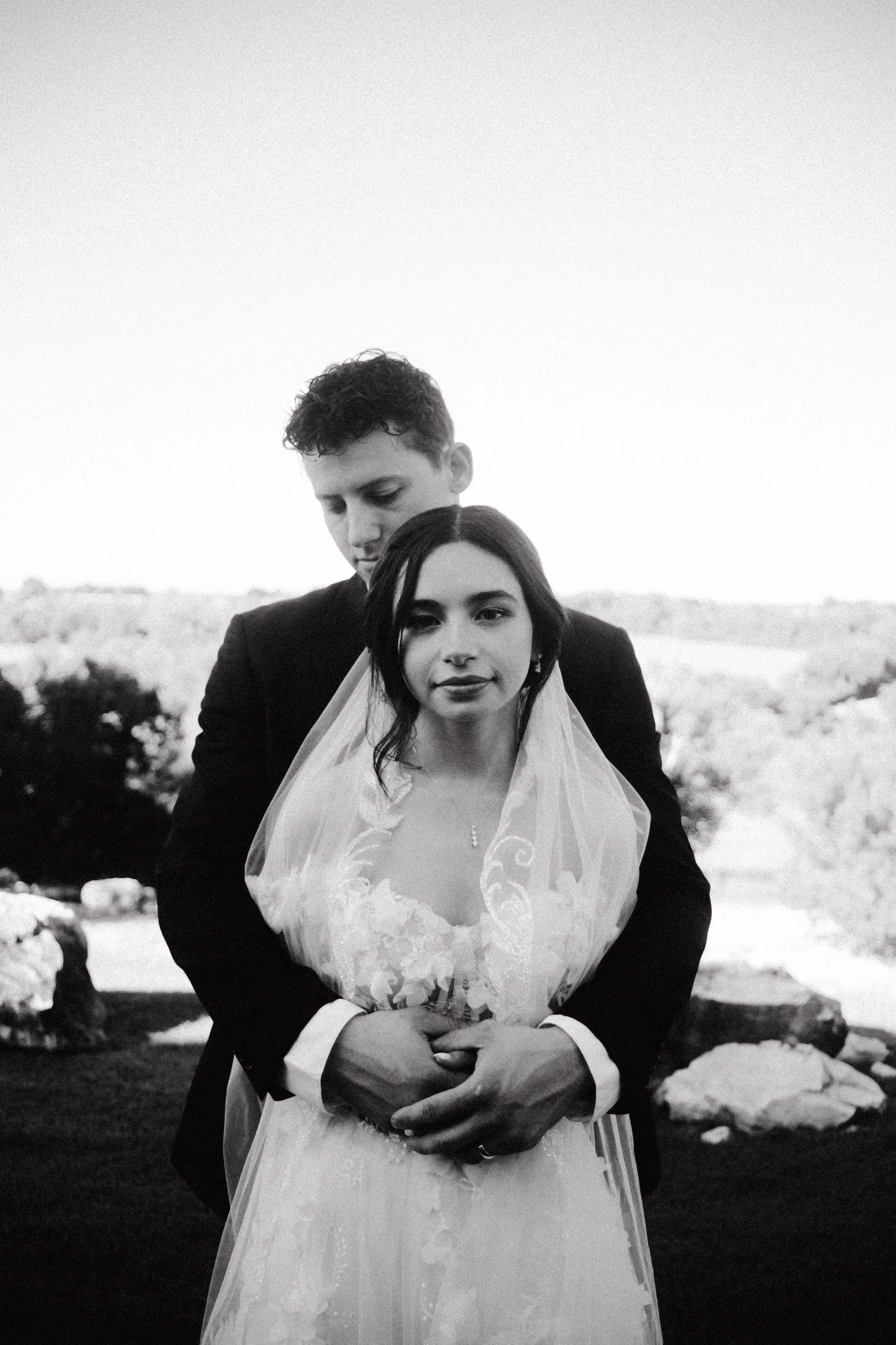 A black and white photo of a bride and groom embracing outdoors, with the groom standing behind the bride, both looking at the camera. The bride is wearing a wedding dress and veil, and the groom is in a dark suit. The background features a landscape with rocks, grass, and trees.
