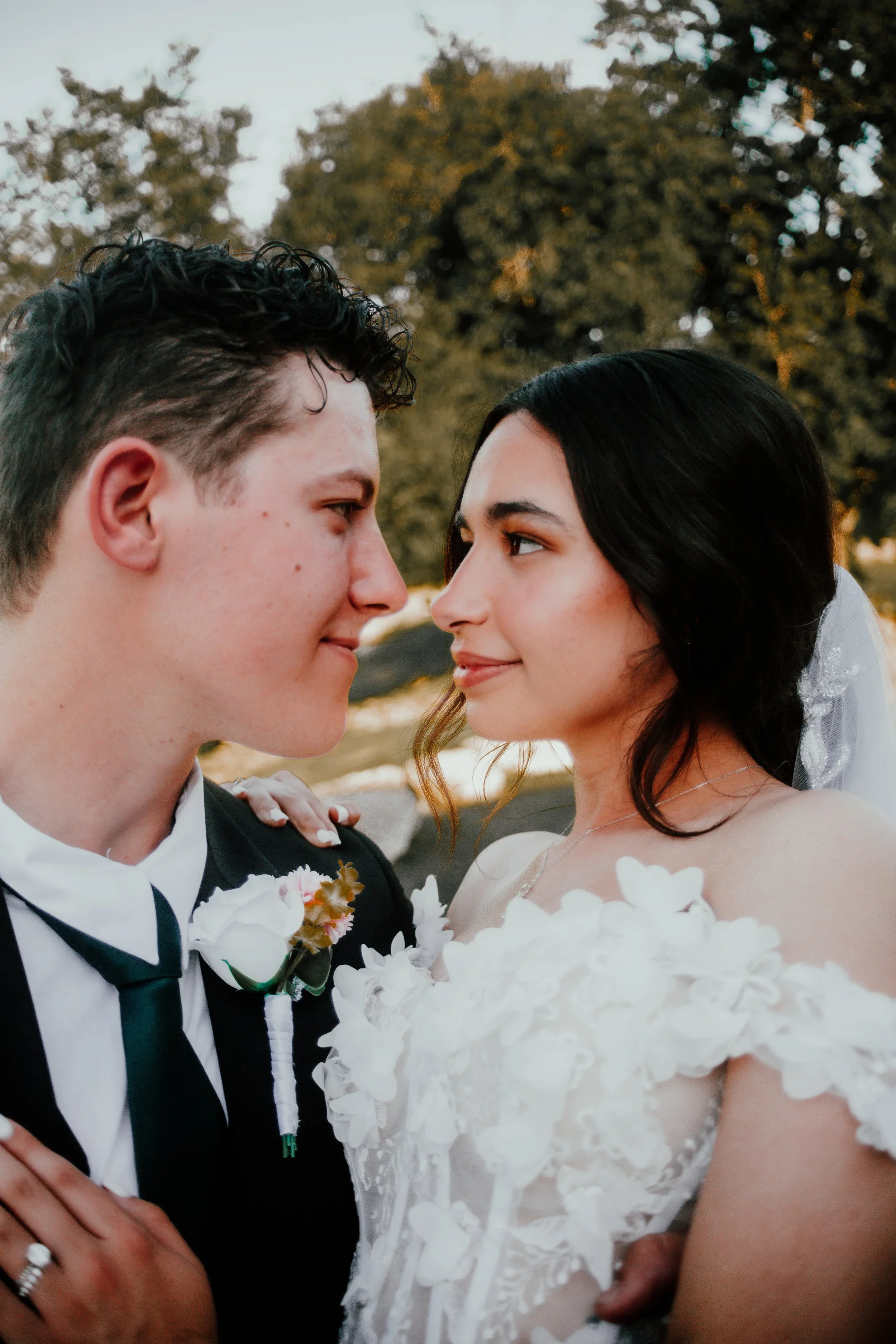 A close-up of a newlywed couple looking at each other, dressed in wedding attire, outdoors with trees in the background.