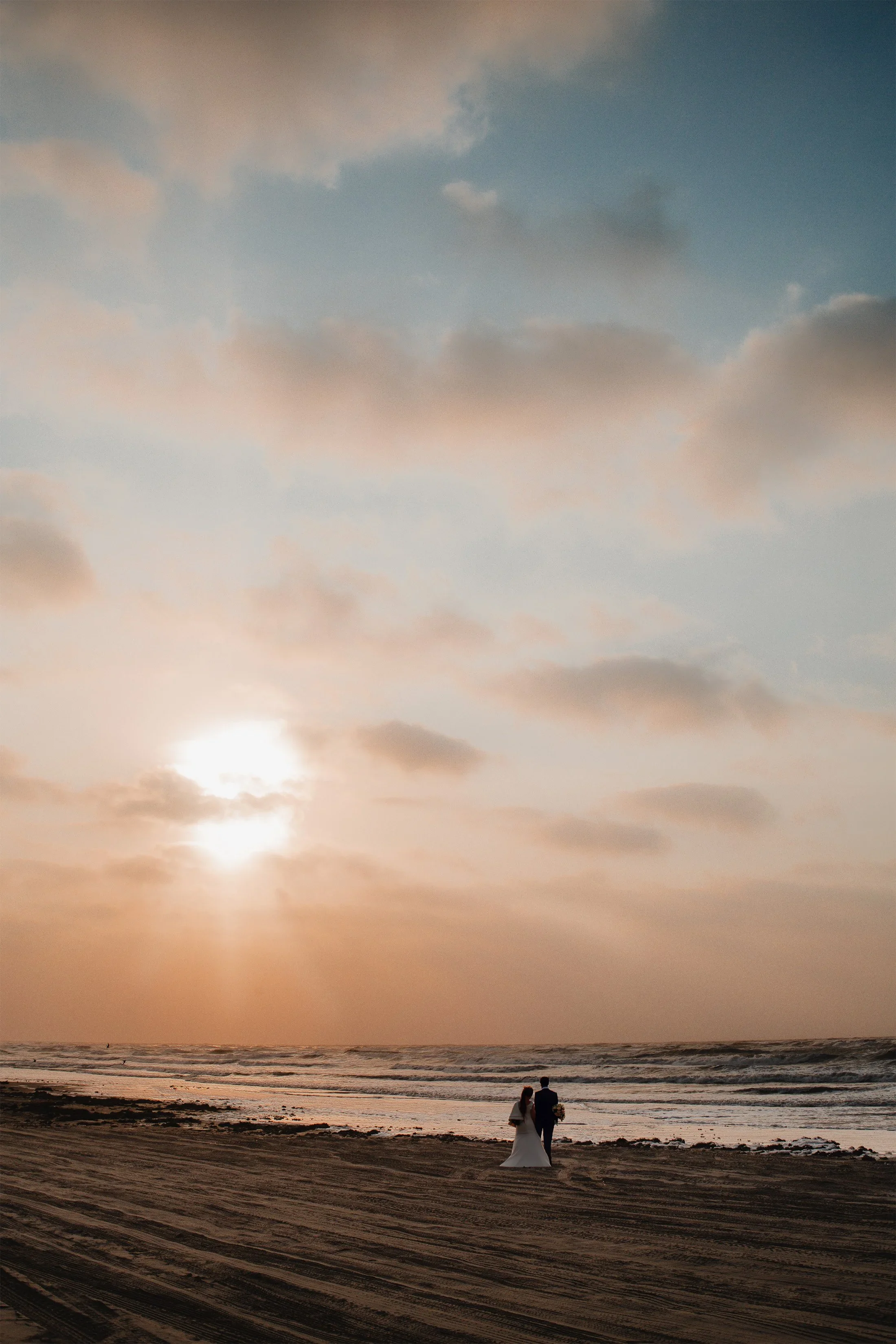 A couple dressed in wedding attire walking along the seashore at sunset, with the sky filled with soft clouds and the sun low in the horizon.