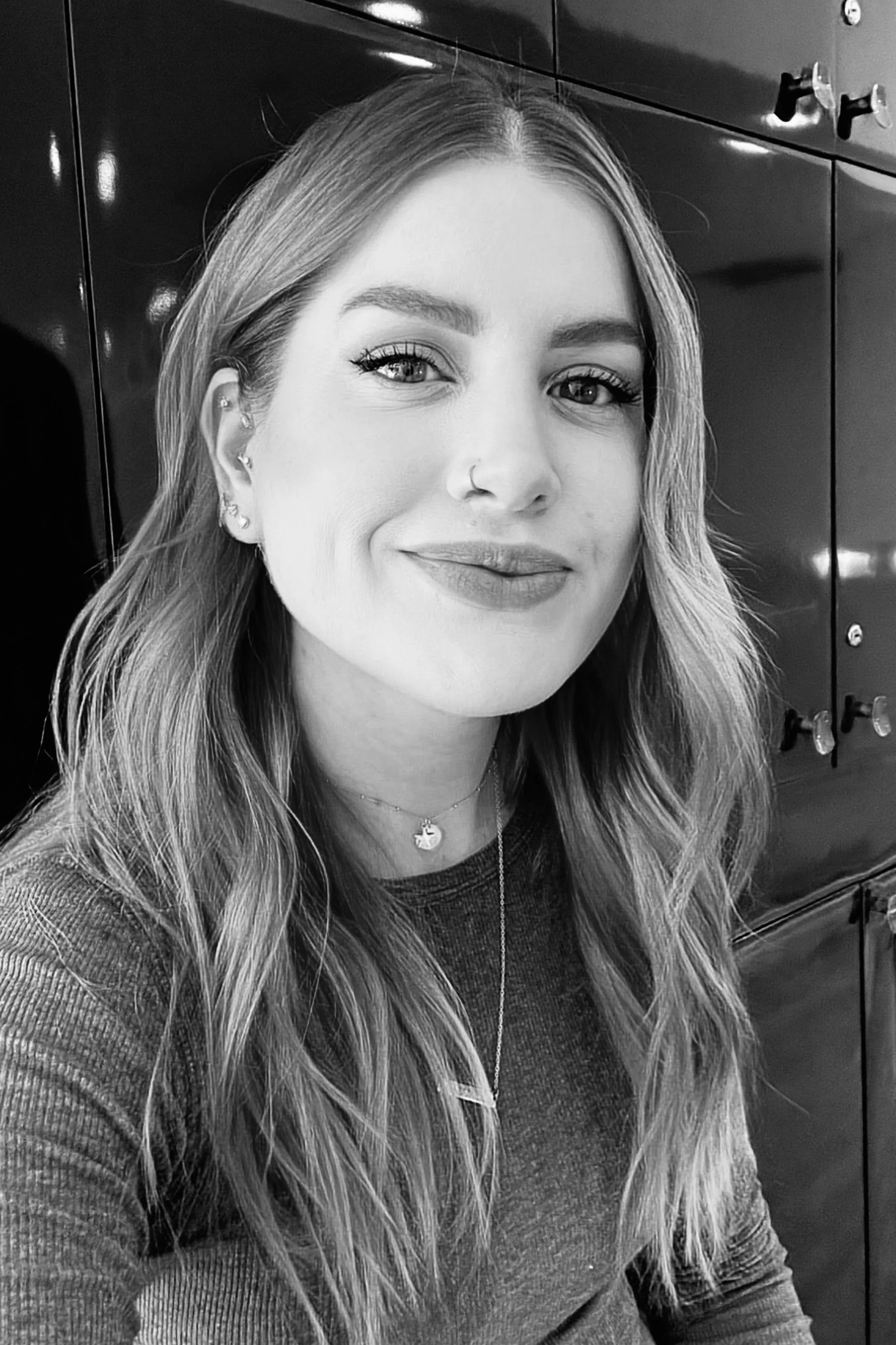 A young woman with long wavy hair, wearing a dark long-sleeve top, jewelry including earrings, necklaces, and a nose ring, smiling in front of a dark, shiny background with lockers.