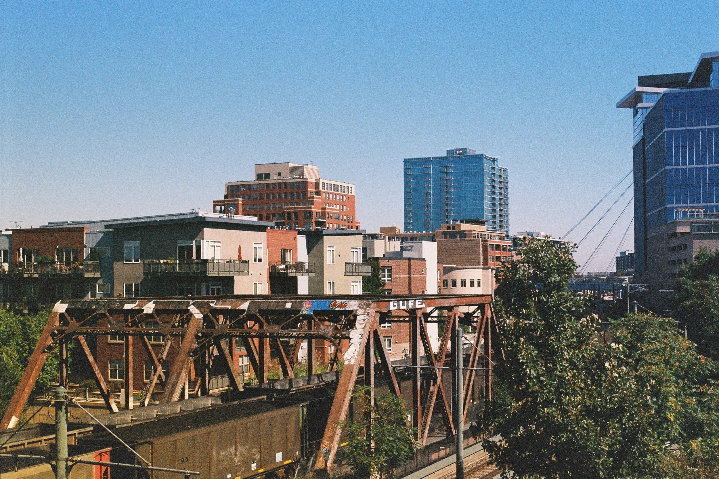 Cityscape with modern high-rise buildings and a rusty train bridge in the foreground under a clear blue sky.