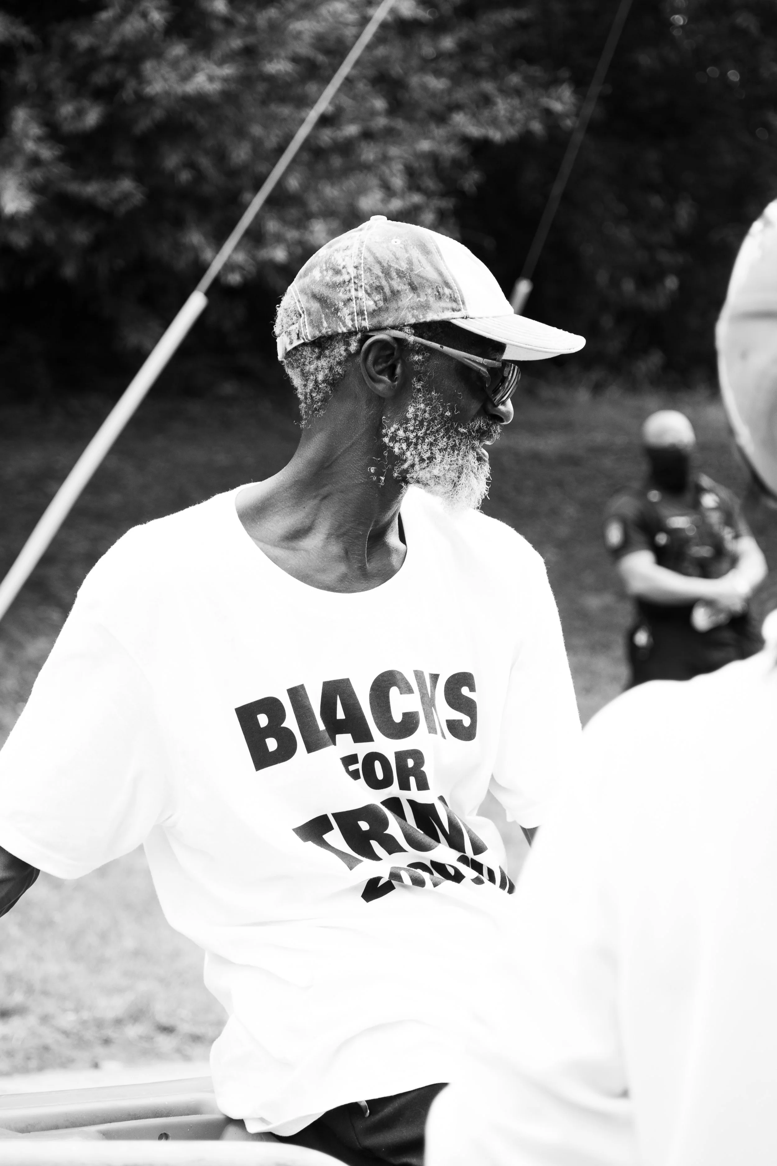 A Black man with white hair and beard, wearing sunglasses, a cap, and a T-shirt that reads 'BLACK IS FOR TRUMP.' He is standing outdoors near a police officer in uniform.