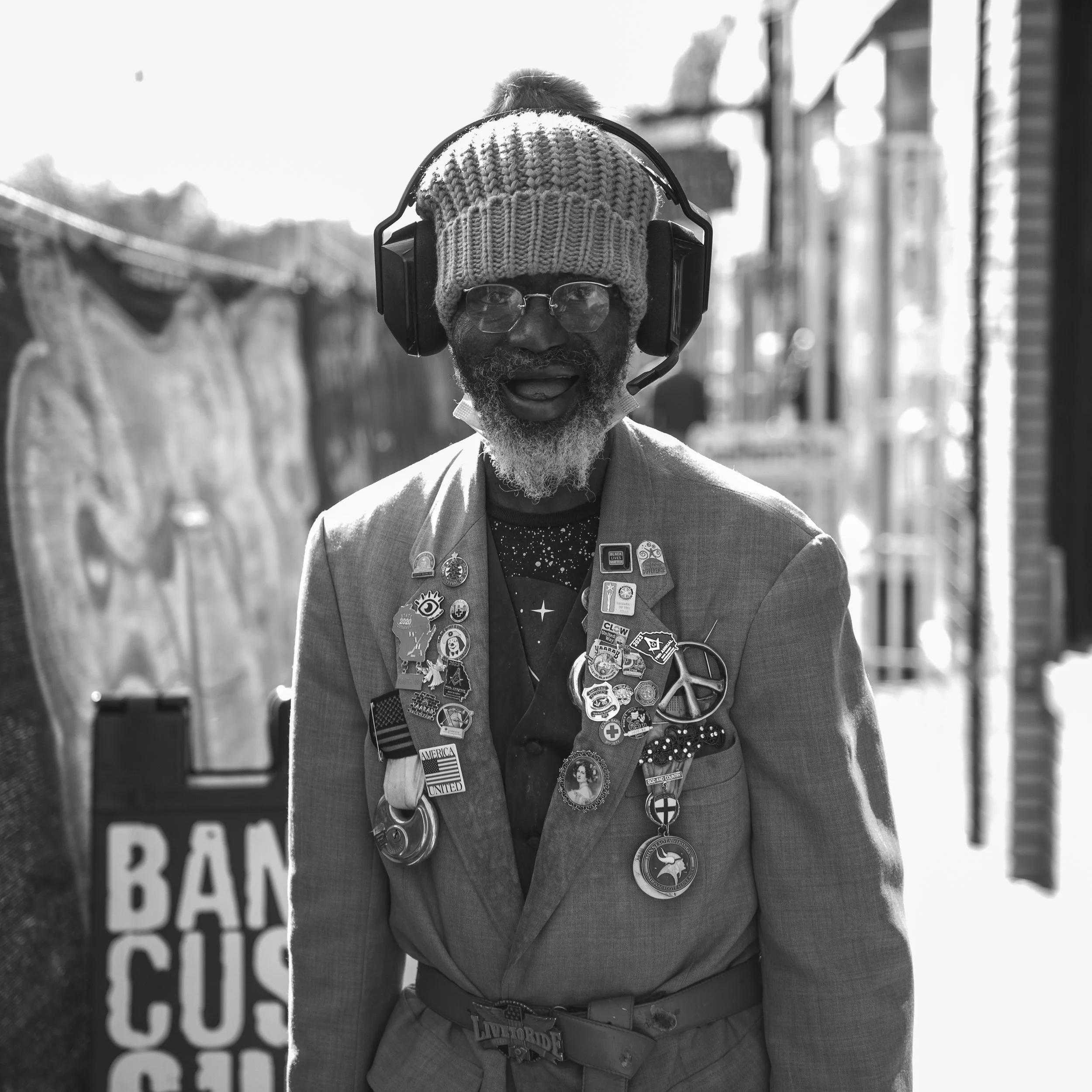 An elderly man with a beard wearing a knit cap, glasses, and a suit jacket covered with various pins and badges, standing outdoors with a sign that reads 'BAN CUSS.'