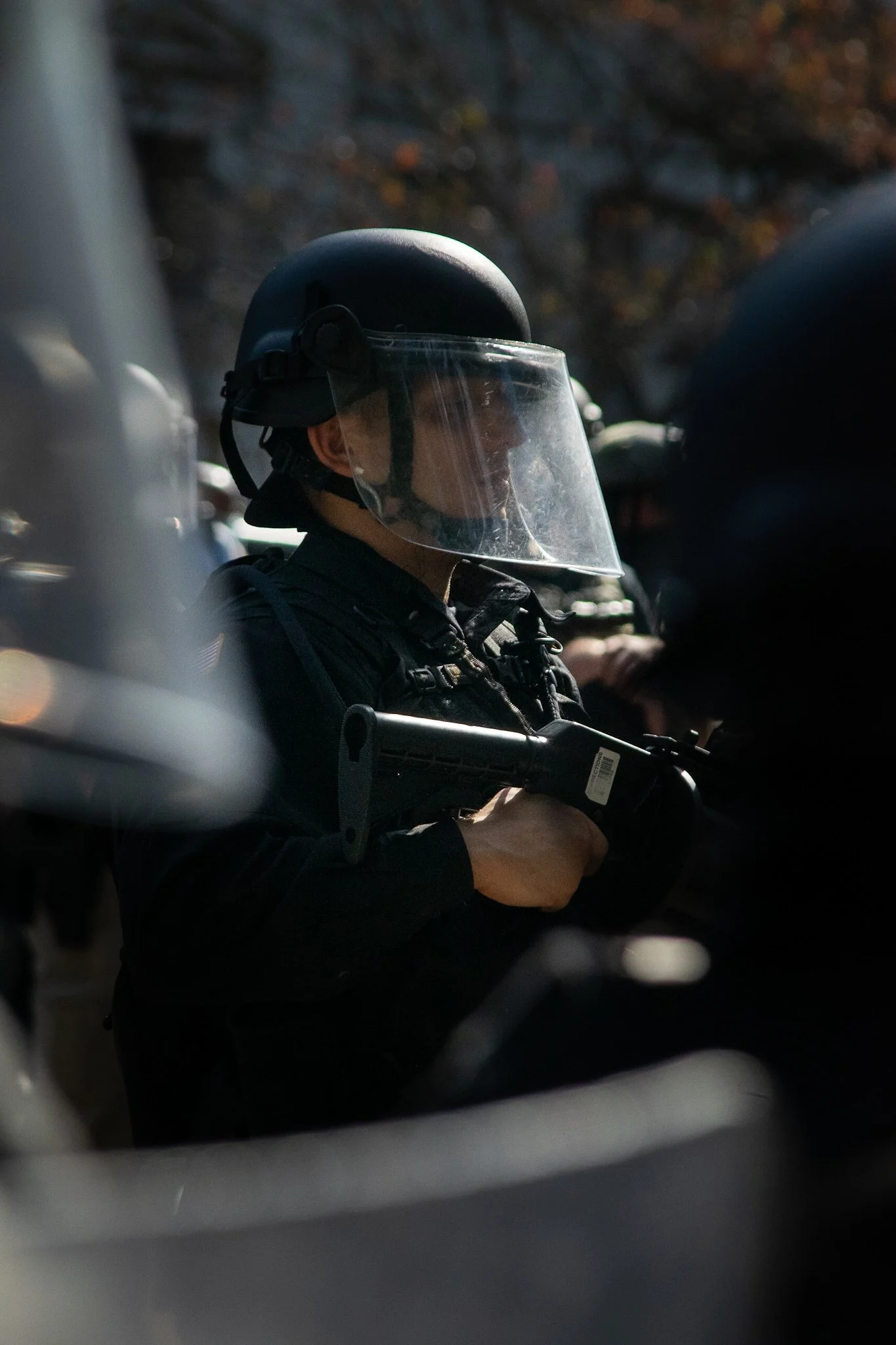 Police officer in tactical gear and helmet with face shield, holding a baton or weapon, during daylight, with other officers in background.