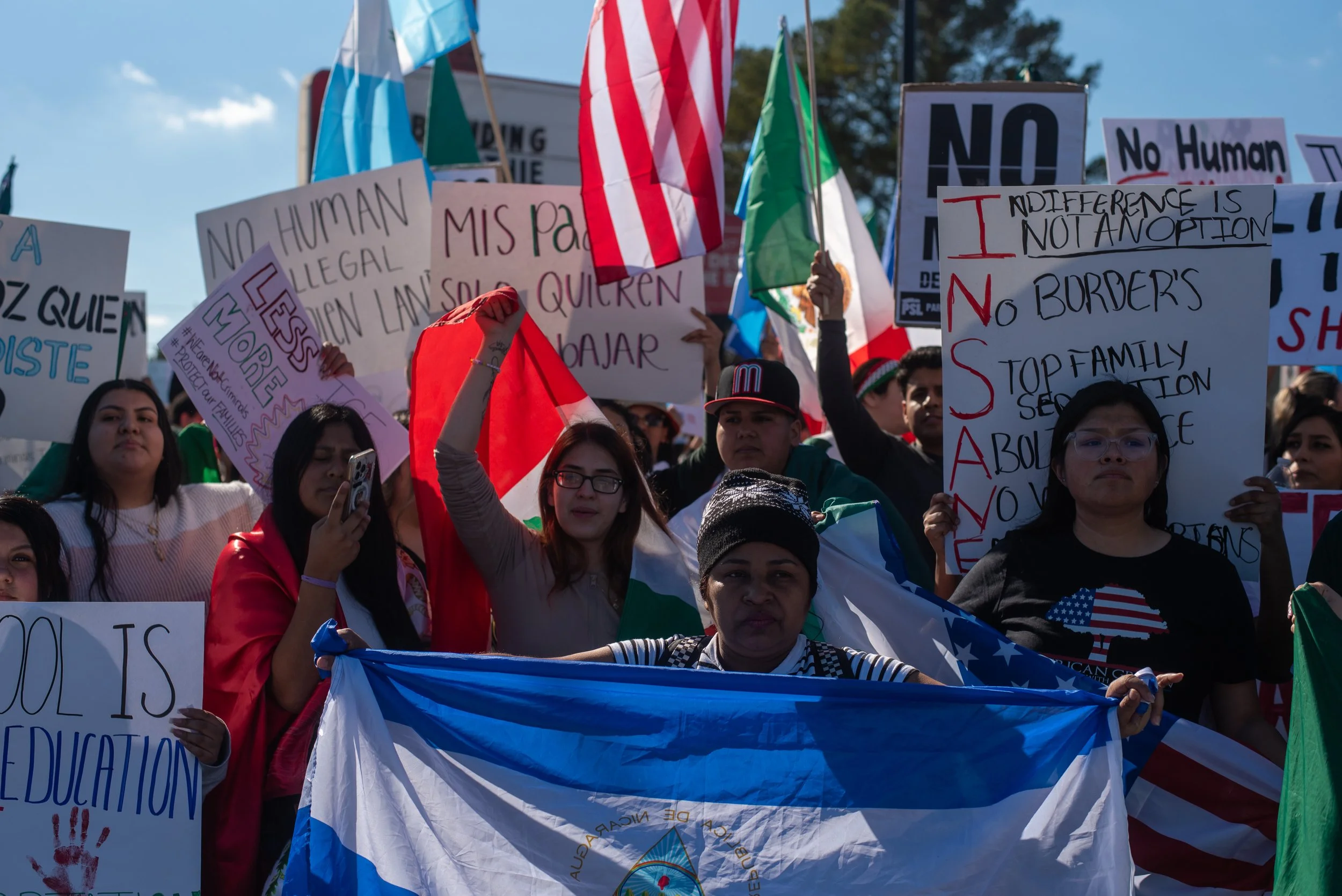 A diverse group of people at a protest holding flags and signs, including a Guatemalan flag and an American flag. The signs display messages related to immigration, border policies, and human rights.