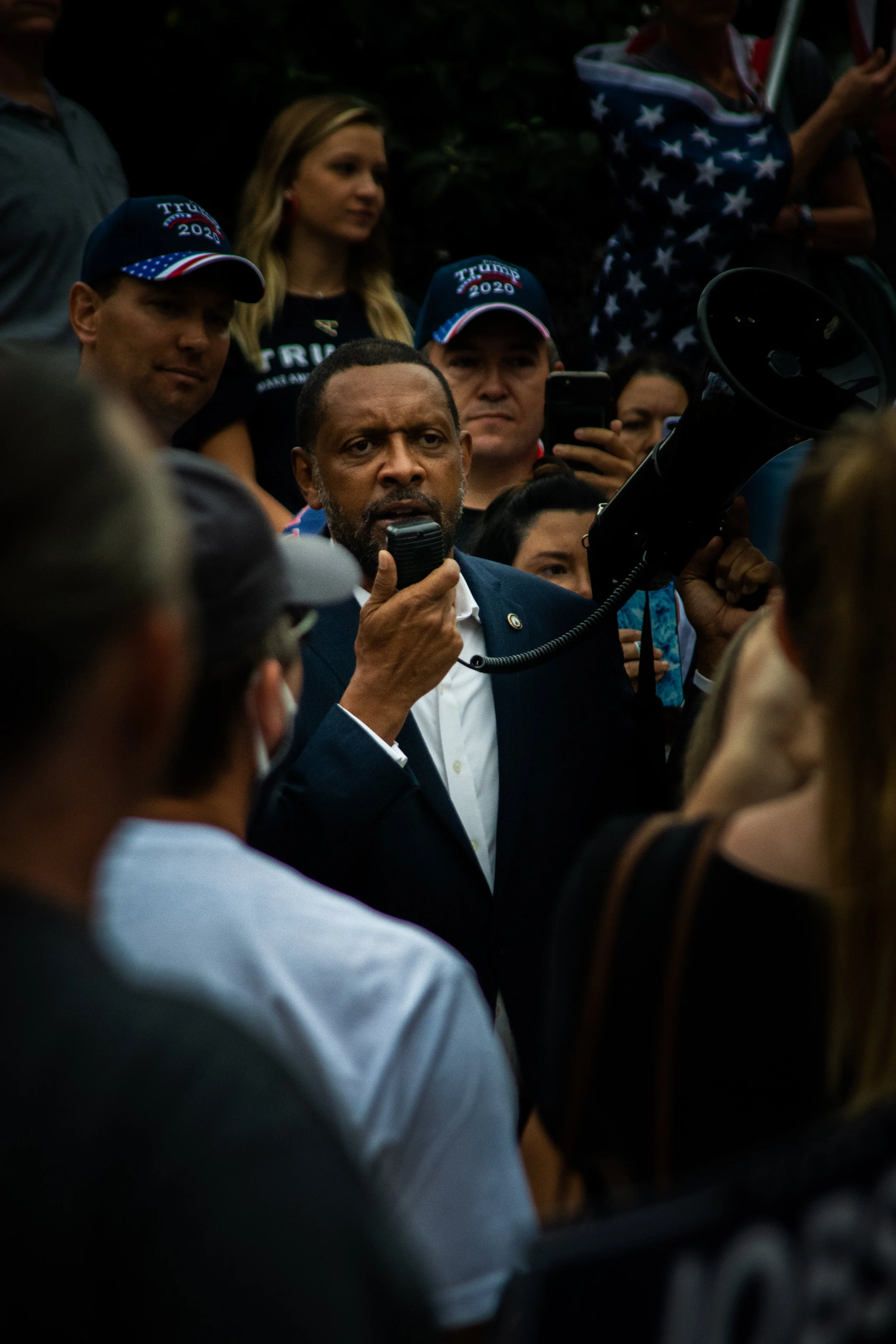 A man speaking through a megaphone at a political rally, surrounded by a crowd of supporters, some wearing 'Trump 2020' hats.