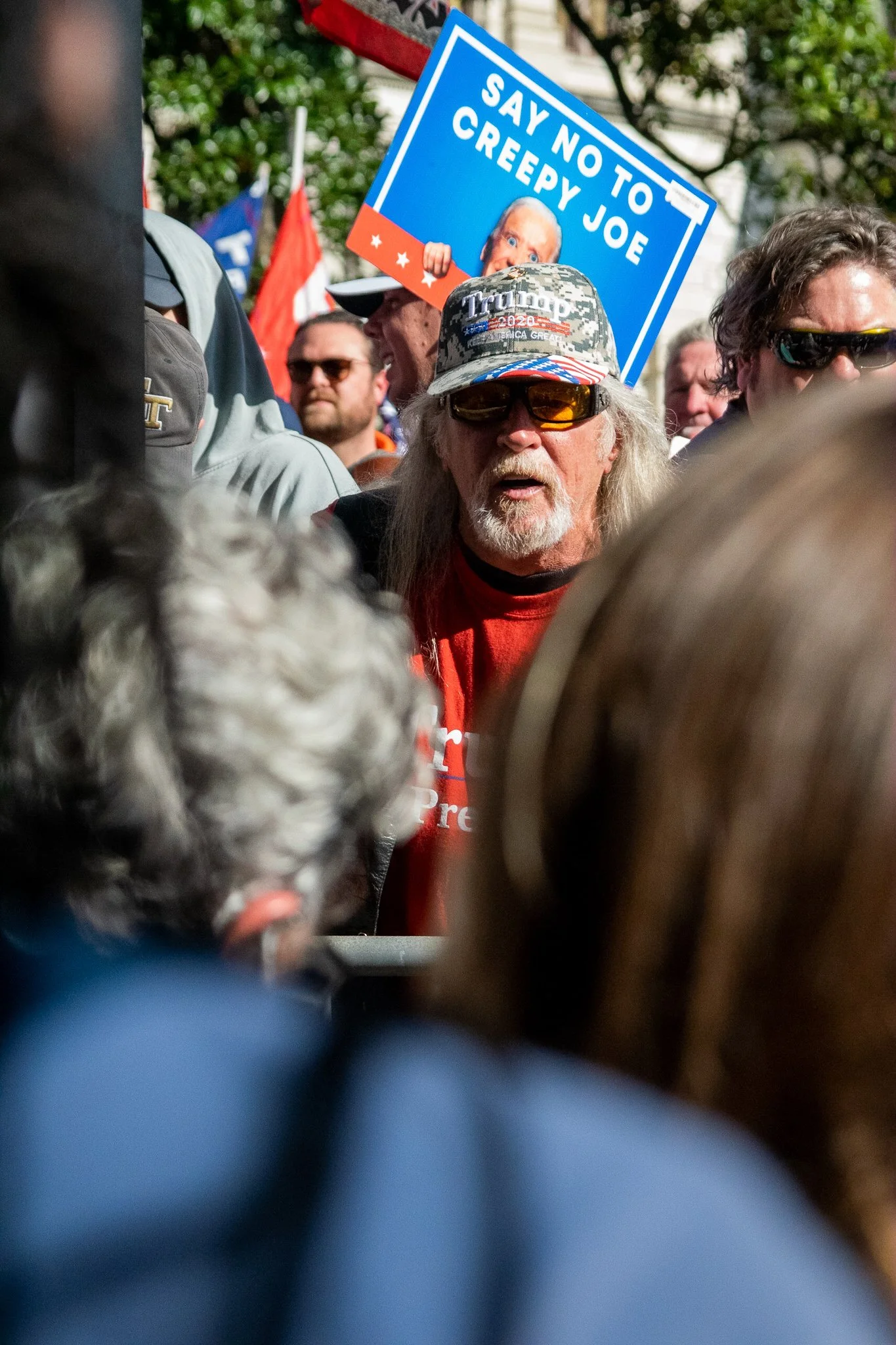 A man wearing a camouflage hat with Trump 2020 embroidered on it, sunglasses, and a red shirt, standing among a crowd of people. In the background, there is a blue sign that reads "Say No to Creepy Joe" with an image of Joe Biden's face.