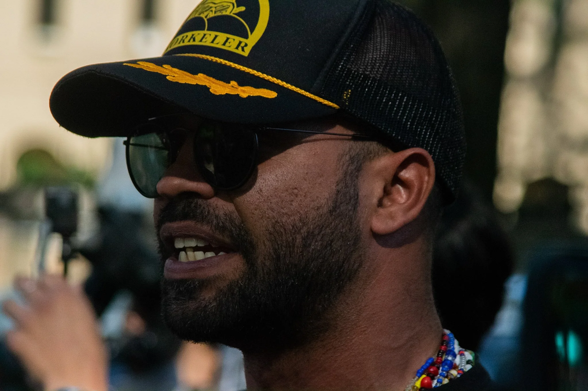 A man wearing a baseball cap with yellow embroidery, sunglasses, and colorful beaded necklaces. The cap has some yellow text and a logo on it.
