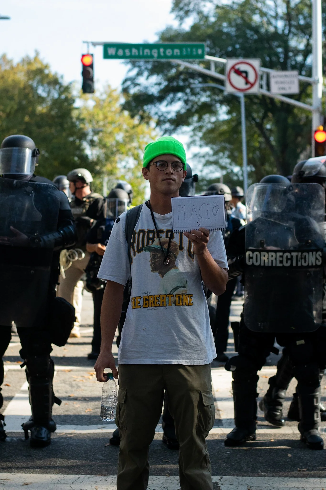 A young man wearing a bright green beanie and glasses holding a sign that reads 'PEACE' with a heart symbol, standing in front of police officers in riot gear during a protest at an intersection.