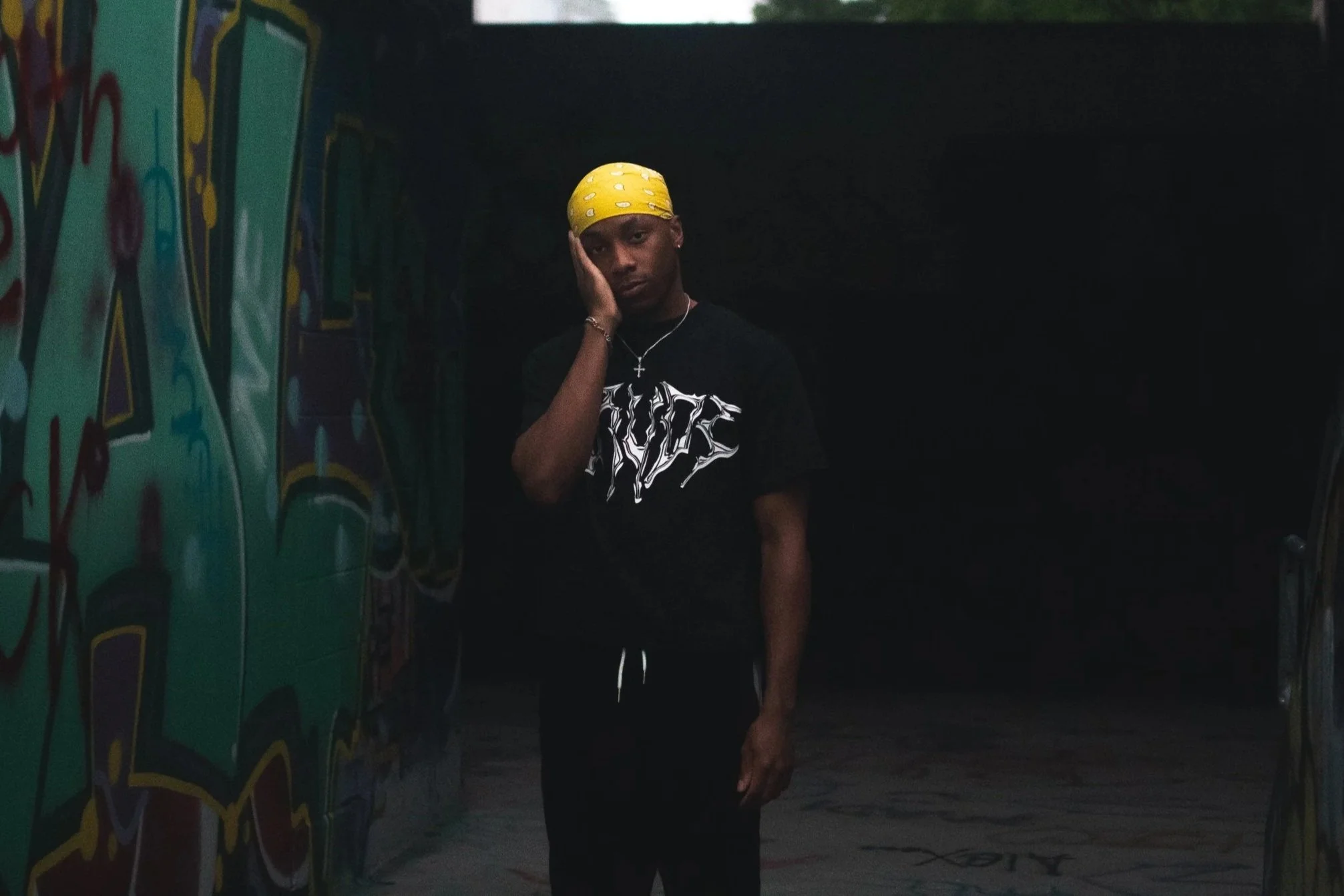 Young man with a yellow bandana, wearing a black T-shirt, standing in front of graffiti-covered wall in a dark setting, looking at the camera.