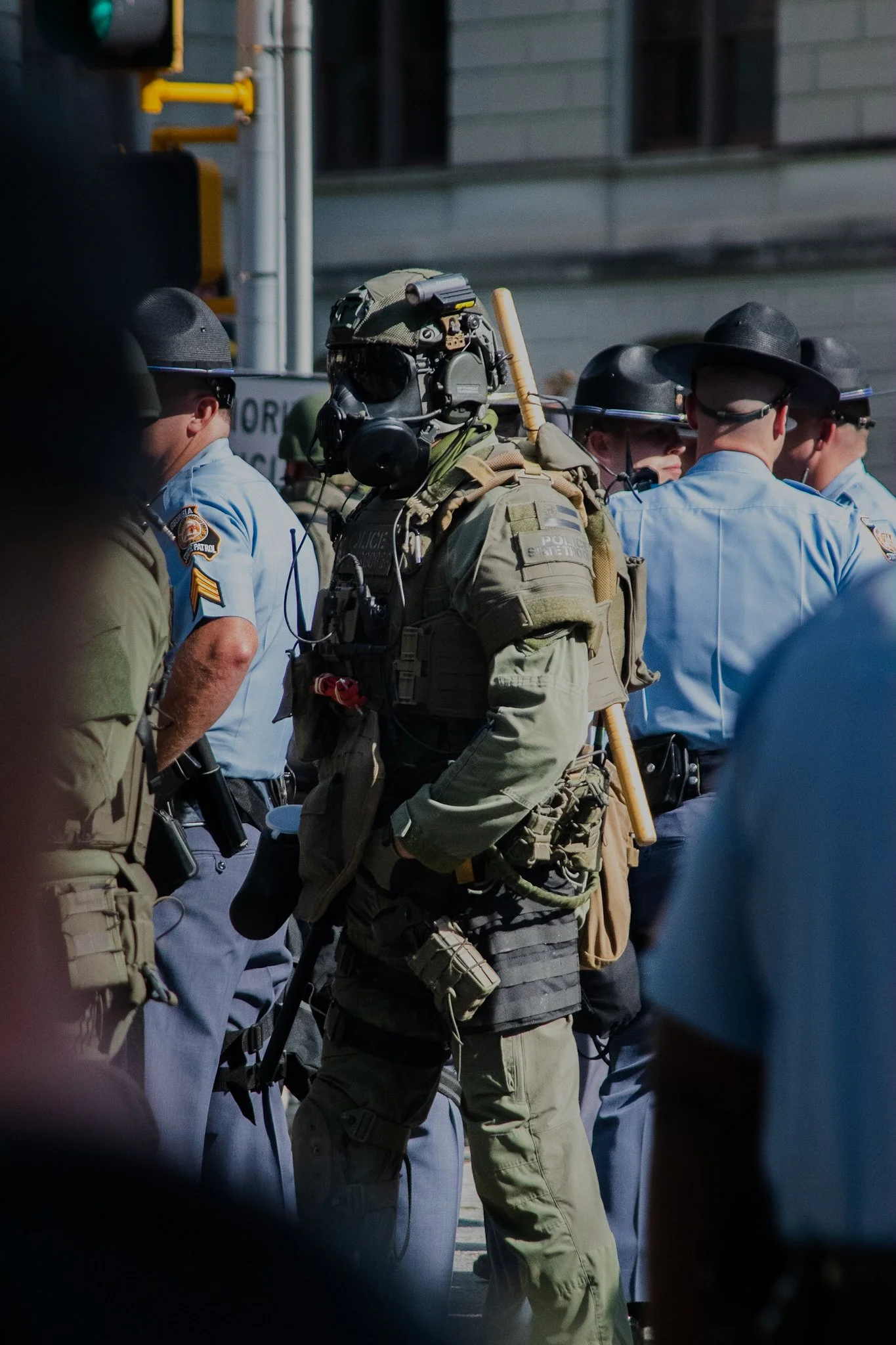 Police officers and a heavily armed tactical officer standing in a crowd, wearing tactical gear, helmets, and sunglasses.