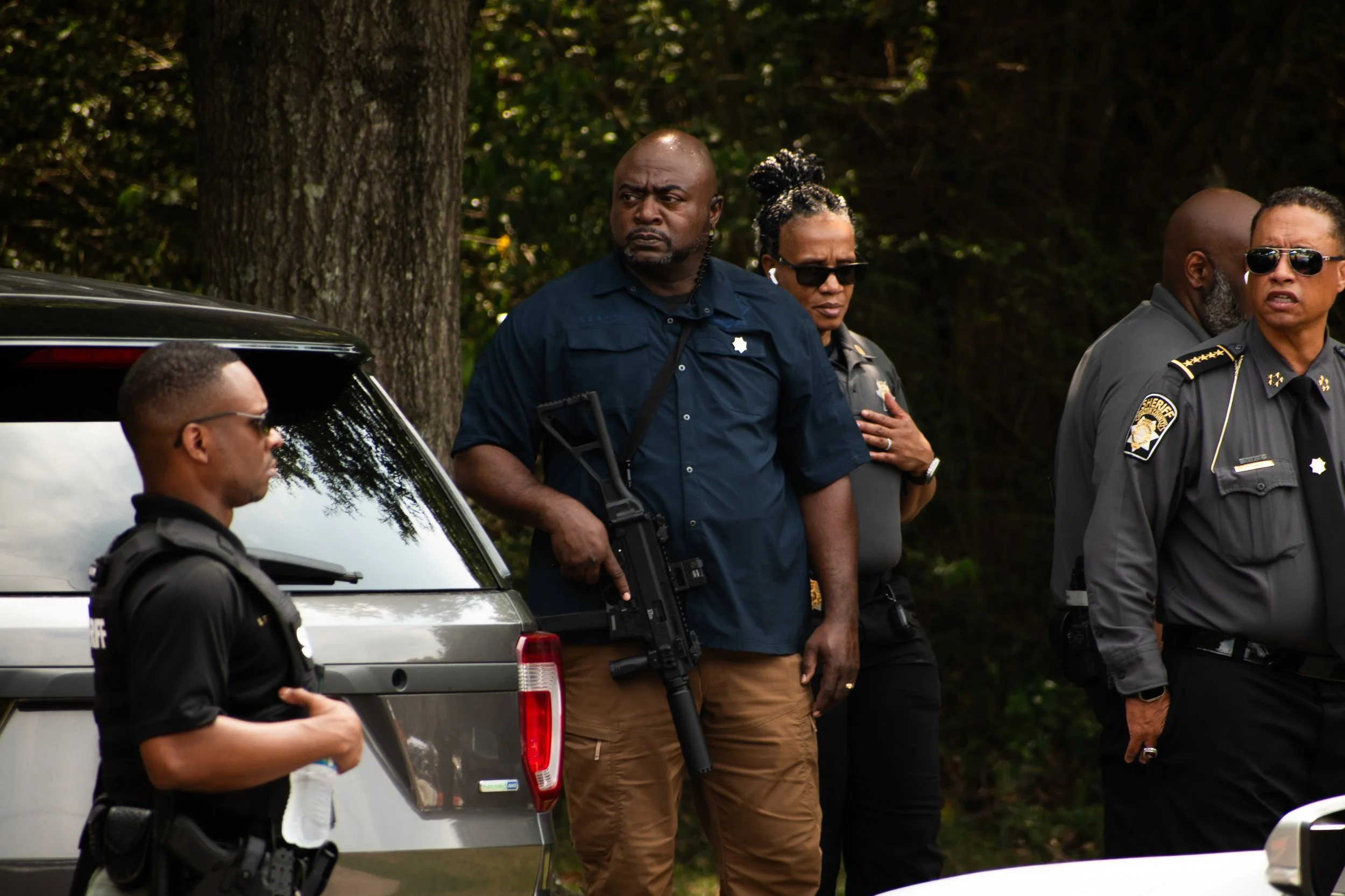Group of police officers standing outdoors beside vehicles, one officer is holding a rifle, some officers are wearing sunglasses and tactical uniforms, in a wooded area.