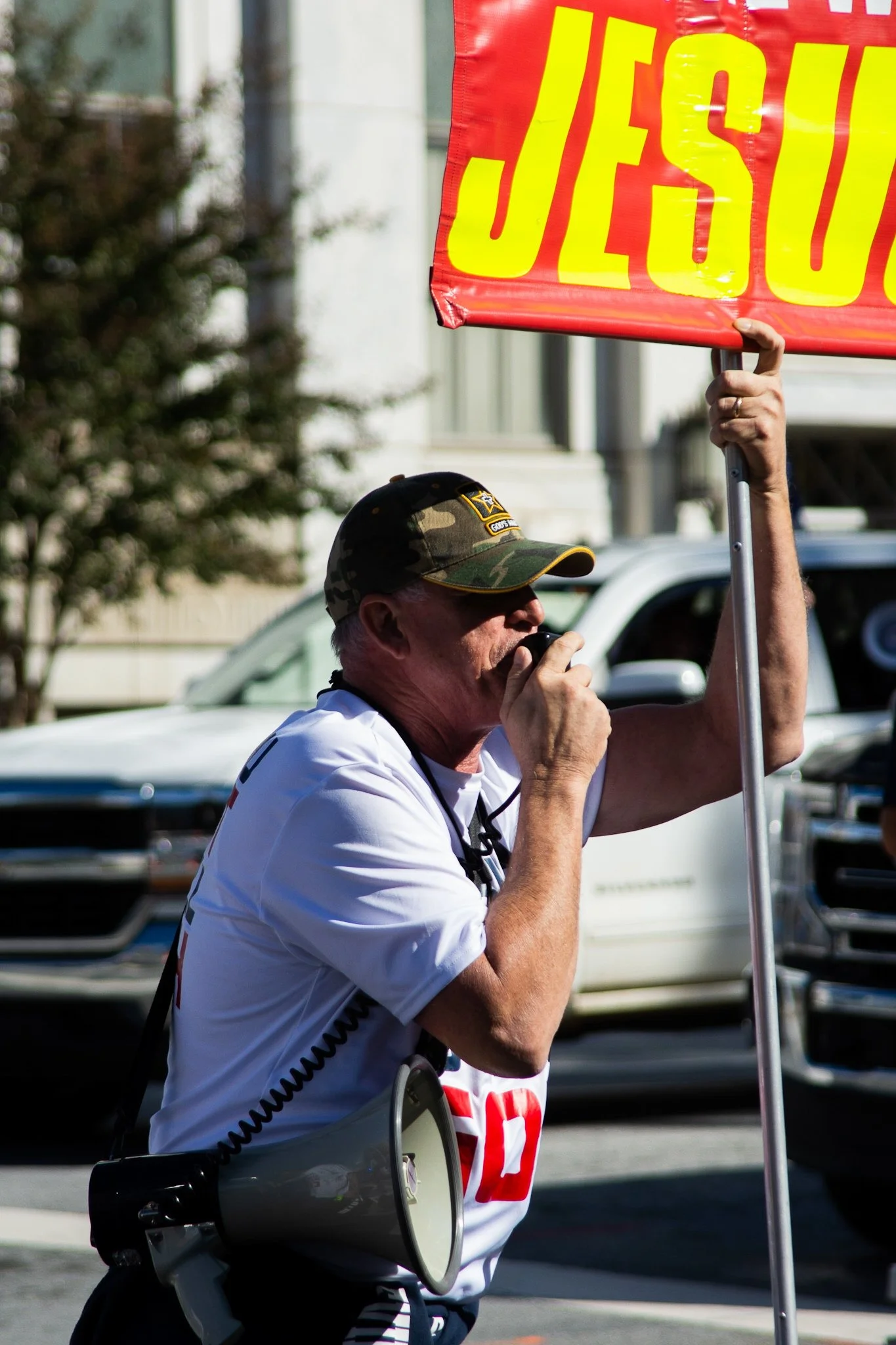 A man wearing a camouflage baseball cap and white T-shirt holding a sign with the word 'JESUS' in yellow letters, speaking into a handheld microphone during a public event or protest.