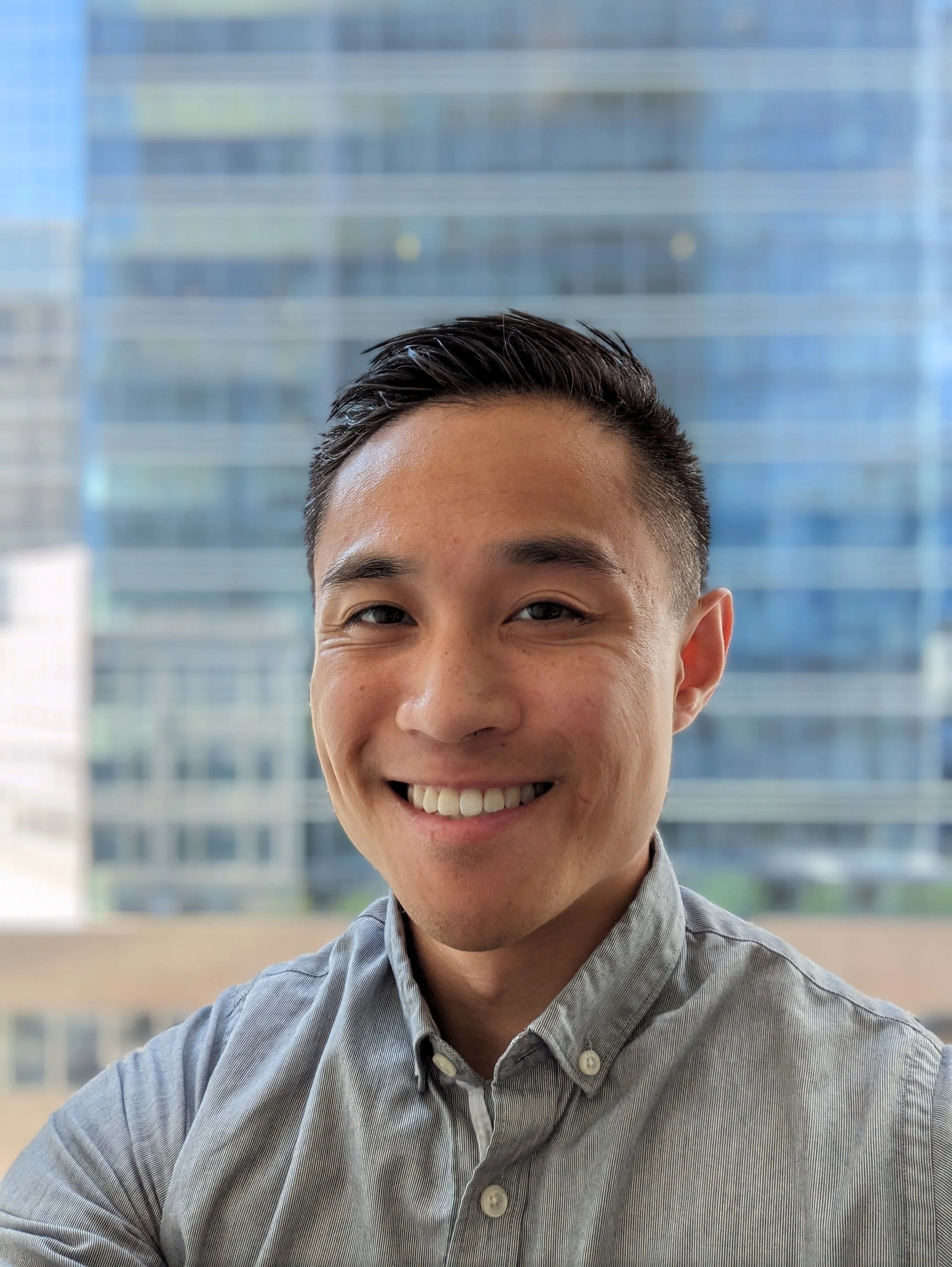 A young man with dark hair smiling, wearing a grey collared shirt, in front of tall office buildings.