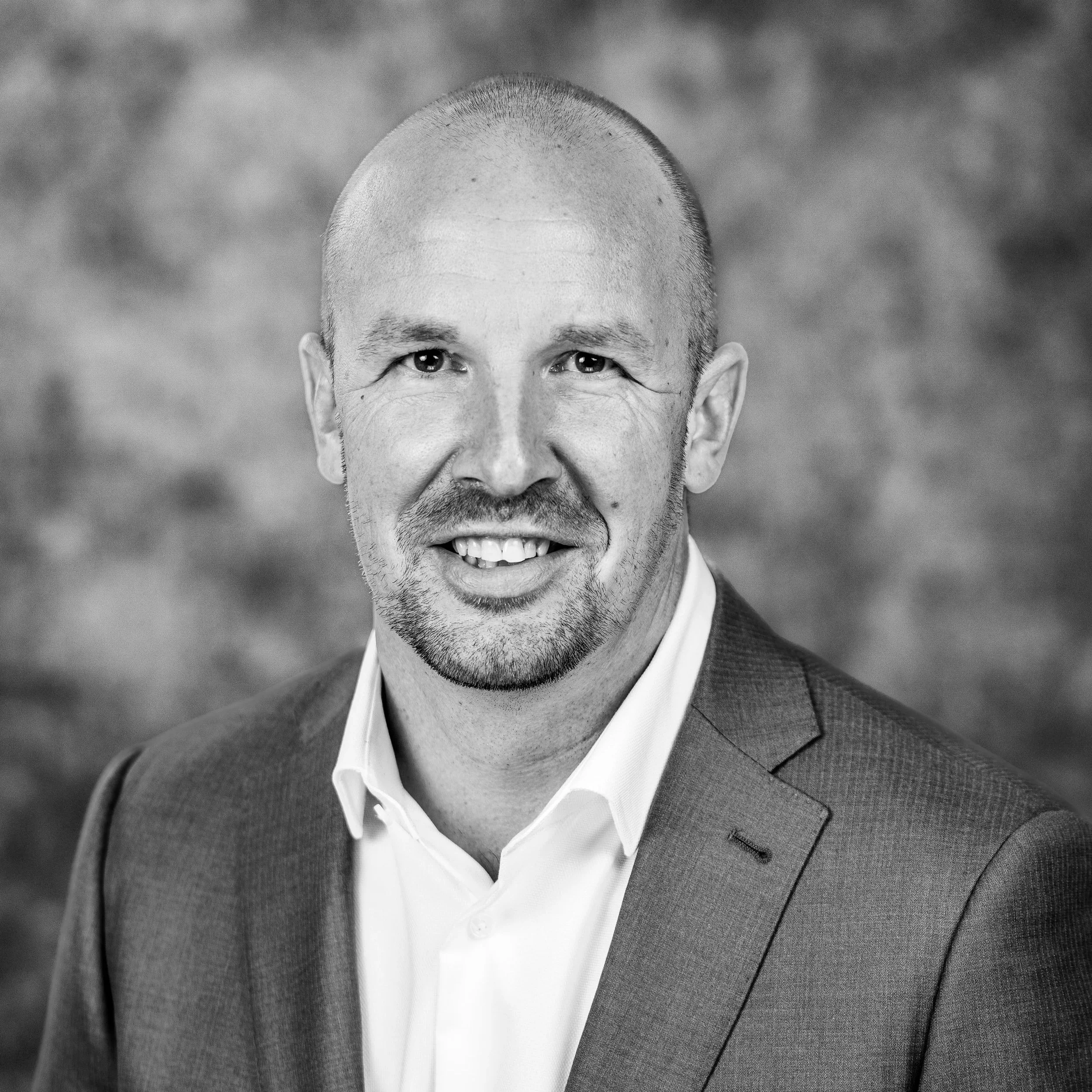 Black and white professional headshot of a smiling man with a shaved head, wearing a suit and dress shirt, standing against a blurred textured background.