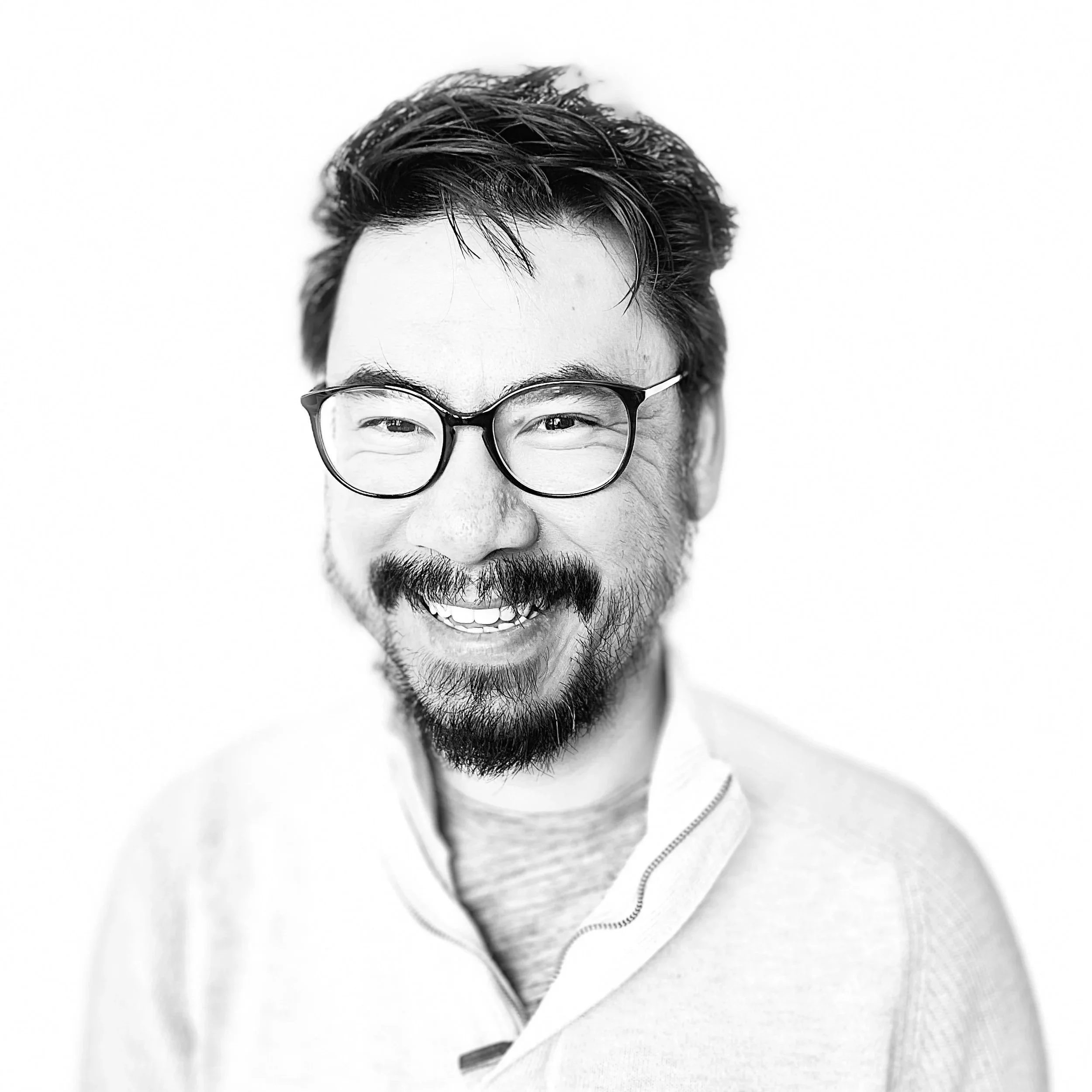 Black and white portrait of a smiling man with glasses, a beard, and short dark hair, wearing a zip-up sweater and a t-shirt, against a plain white background.